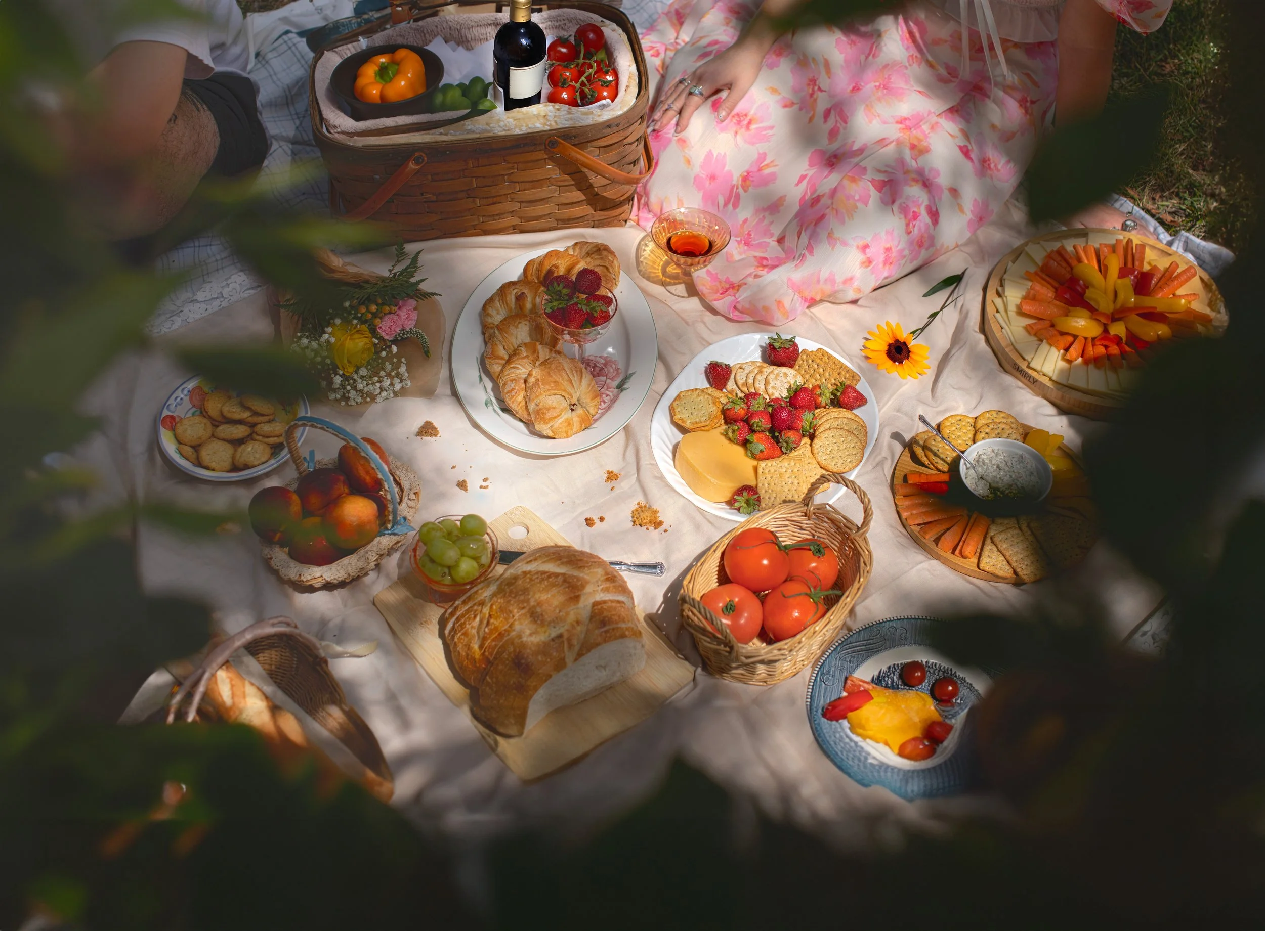A picnic setup with various foods including croissants, strawberries, cheese, crackers, vegetables, grapes, tomatoes, and a variety of cheeses on plates and baskets. There is also a bottle of wine, a glass of rosé, and some flowers on the table.