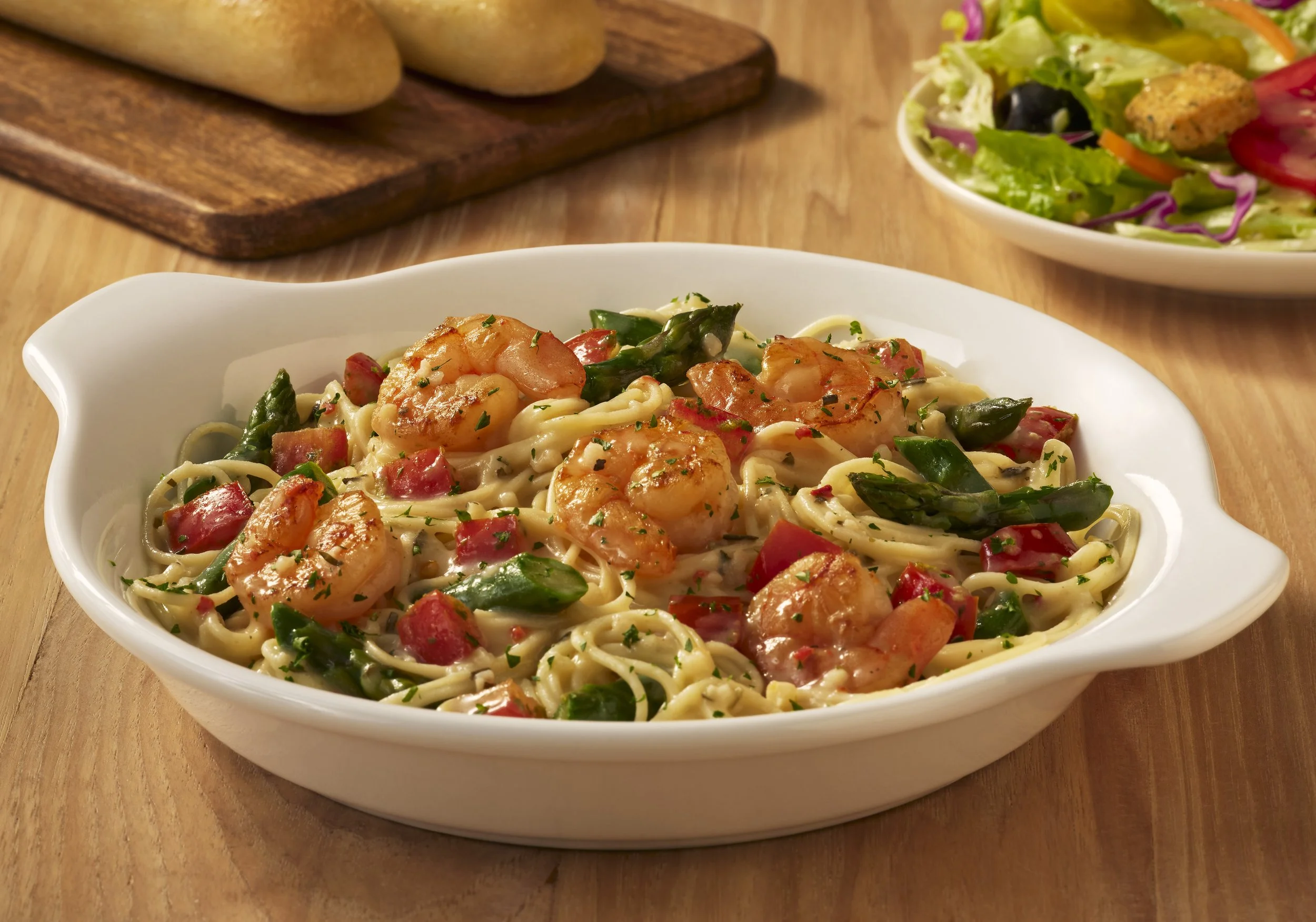 A bowl of pasta with shrimp, green vegetables, and red seasoning, served on a white dish on a wooden table, with a side salad and bread in the background.