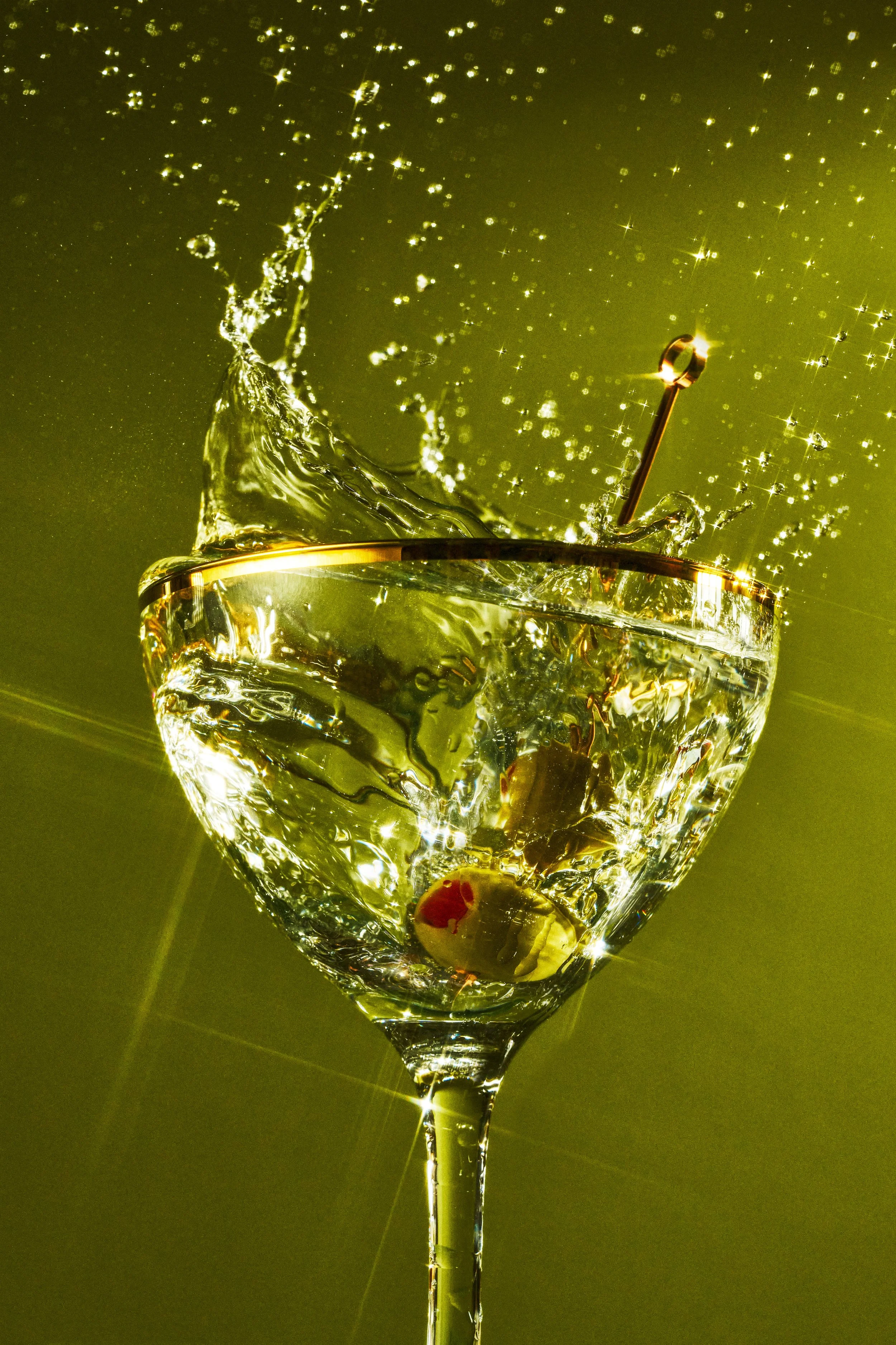 A glass of champagne filled with liquid and a cherry at the bottom, with a drop of champagne falling into the glass creating a splash. The background is greenish with sparkling reflections.