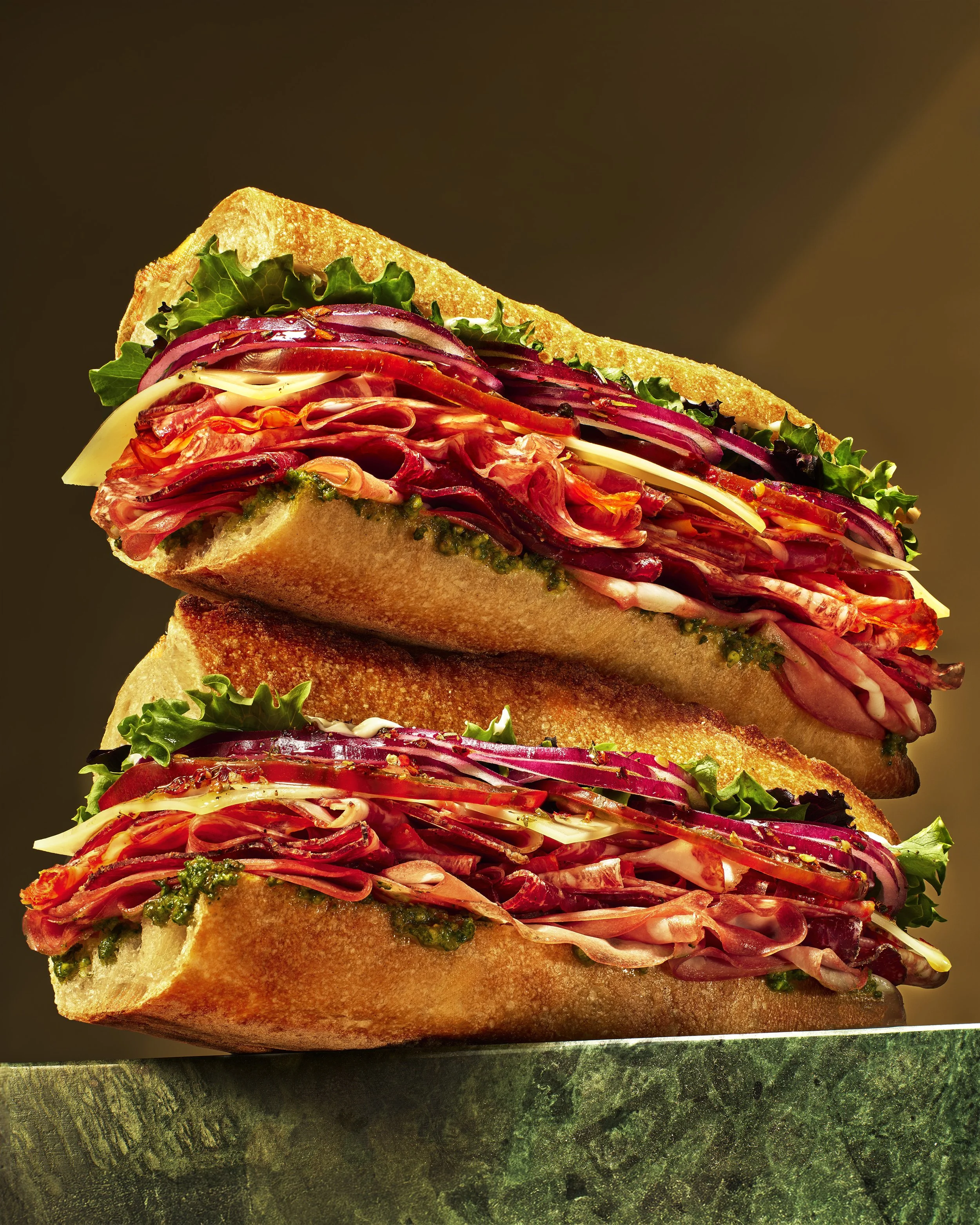 A close-up view of a stacked sandwich with multiple layers of sliced meats, cheeses, red onion, and lettuce on toasted bread with green sauce.
