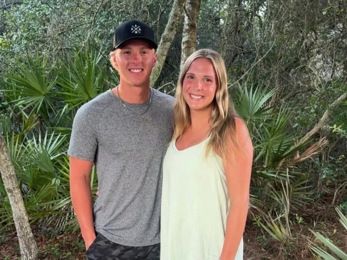 A young man and woman standing outdoors in a wooded area, smiling at the camera.