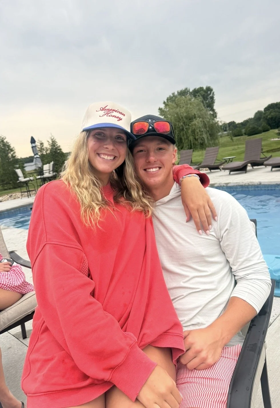 Two young adults, woman and man, smiling and sitting close by a swimming pool in a backyard. The woman wears a white trucker hat with red text, a red sweatshirt, and has long wavy hair. The man wears a white hoodie and red striped shorts, with sunglasses on his head. There are lounge chairs and greenery in the background.