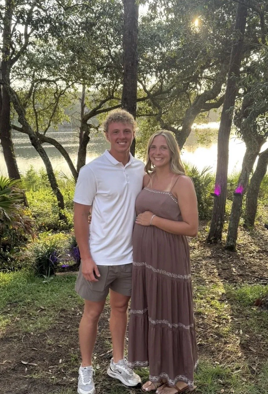 A young man and woman standing outdoors near a lake with trees and sunlight in the background during sunset. The man is wearing a white polo shirt, shorts, and sneakers. The woman is wearing a long, brown strapless dress and sandals. They are smiling and posing together.
