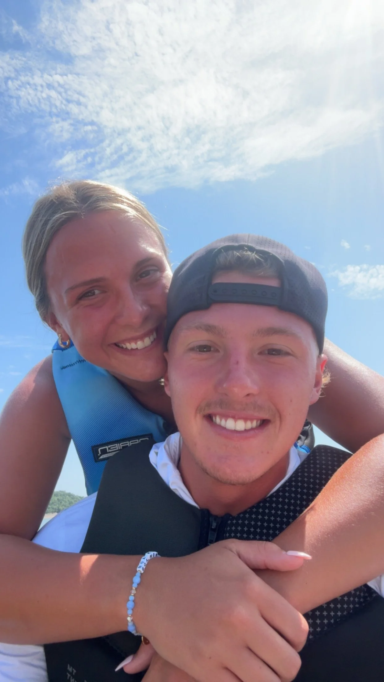 A woman and a young man are smiling closely together in an outdoor setting under a blue sky with clouds, with the woman hugging the young man from behind.