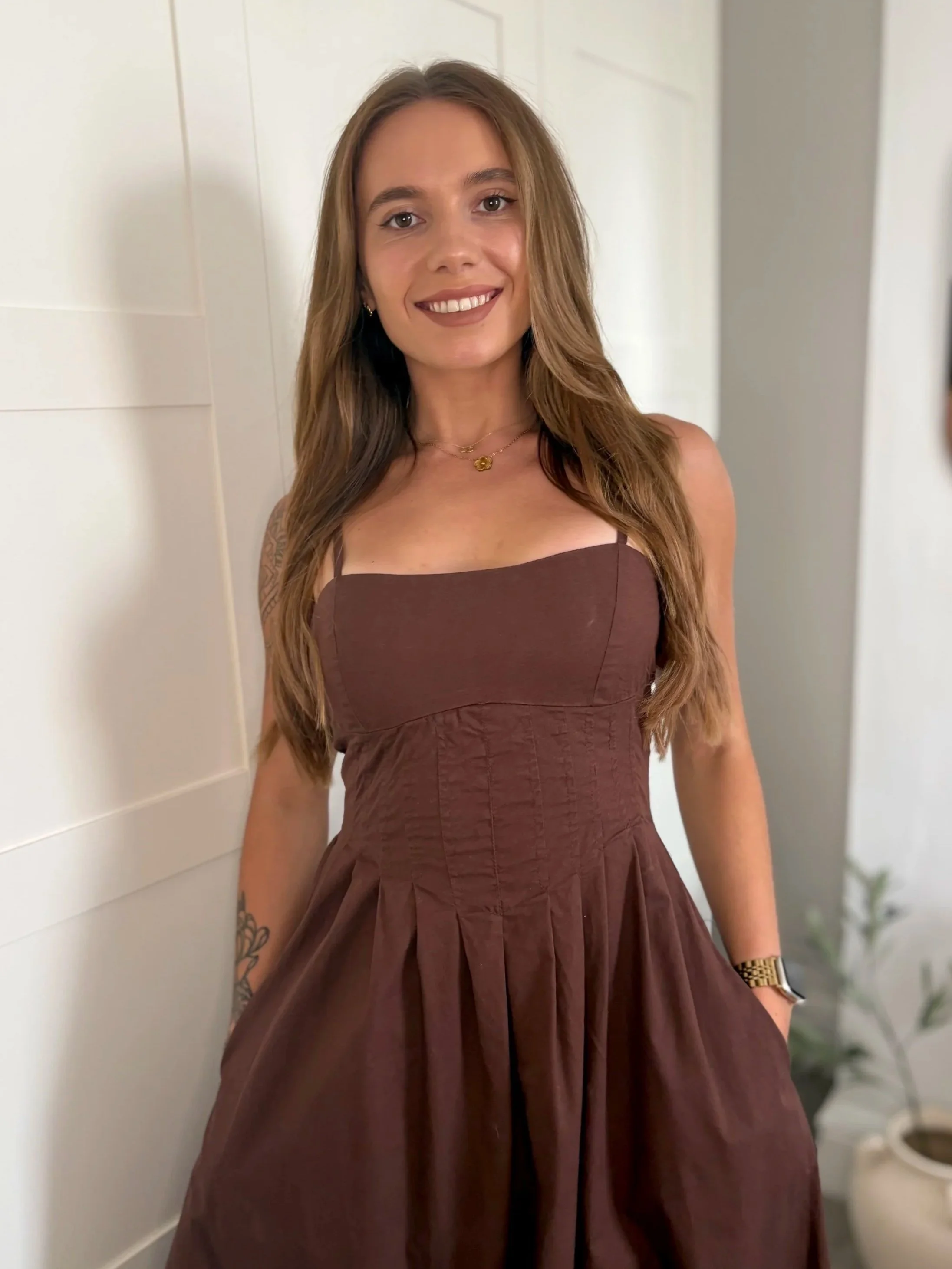 A young woman with long brown hair, wearing a brown dress, standing indoors against a white wall, smiling at the camera.