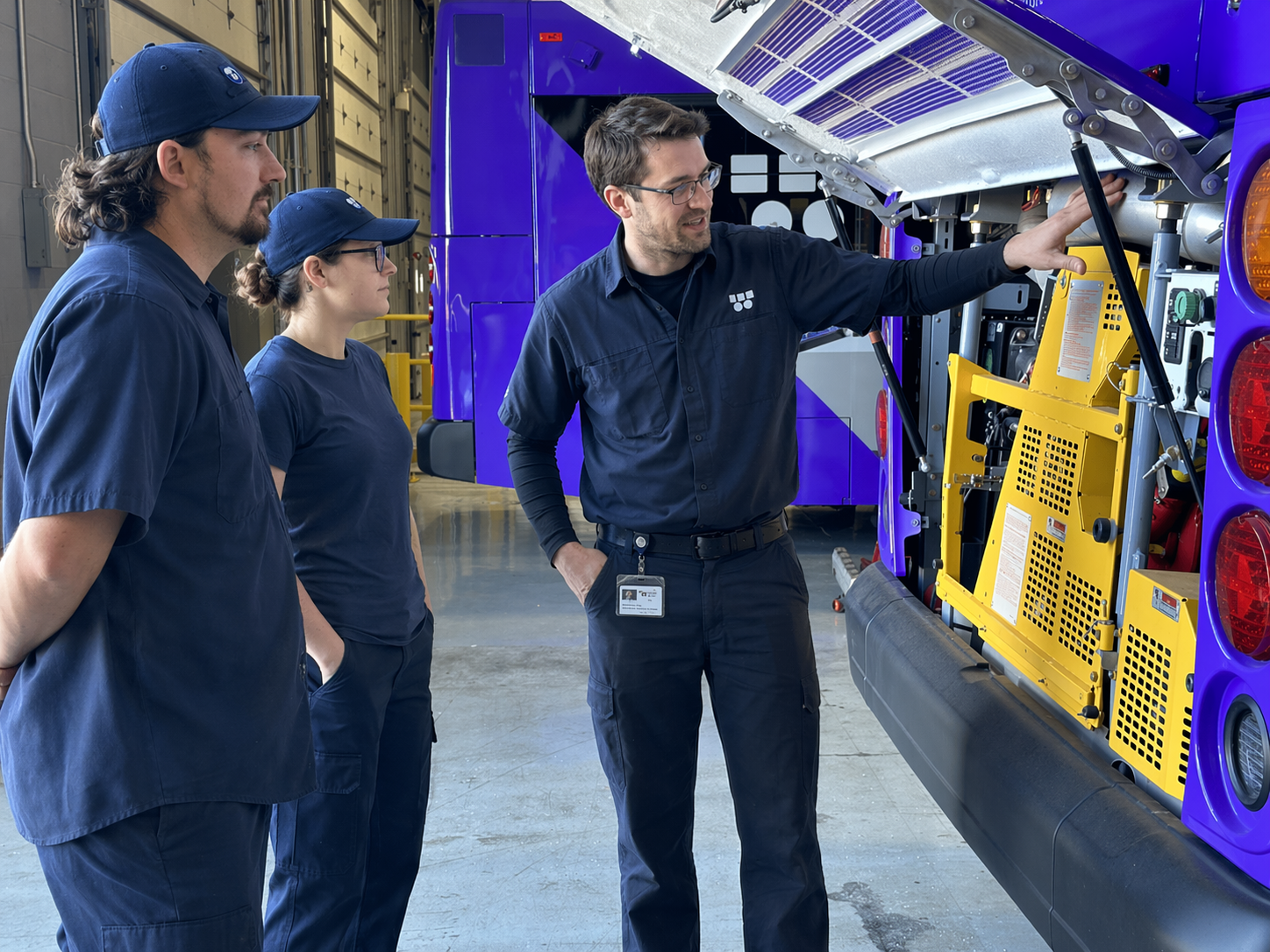 A team of three technicians in navy uniforms and caps inspecting the internal components of a hydrogen bus with an open service panel inside a maintenance garage.