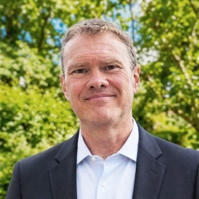 Jim Fouch wearing a dark blazer and white shirt, standing outdoors with green trees in the background.