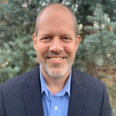 Buford Barr with a beard and short hair, wearing a dark blazer and light blue shirt, standing outdoors with trees in the background.