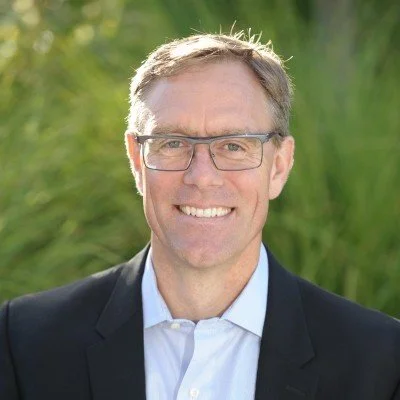 Seth Terry with glasses smiling outdoors, wearing a black blazer and white shirt, with green foliage in the background.