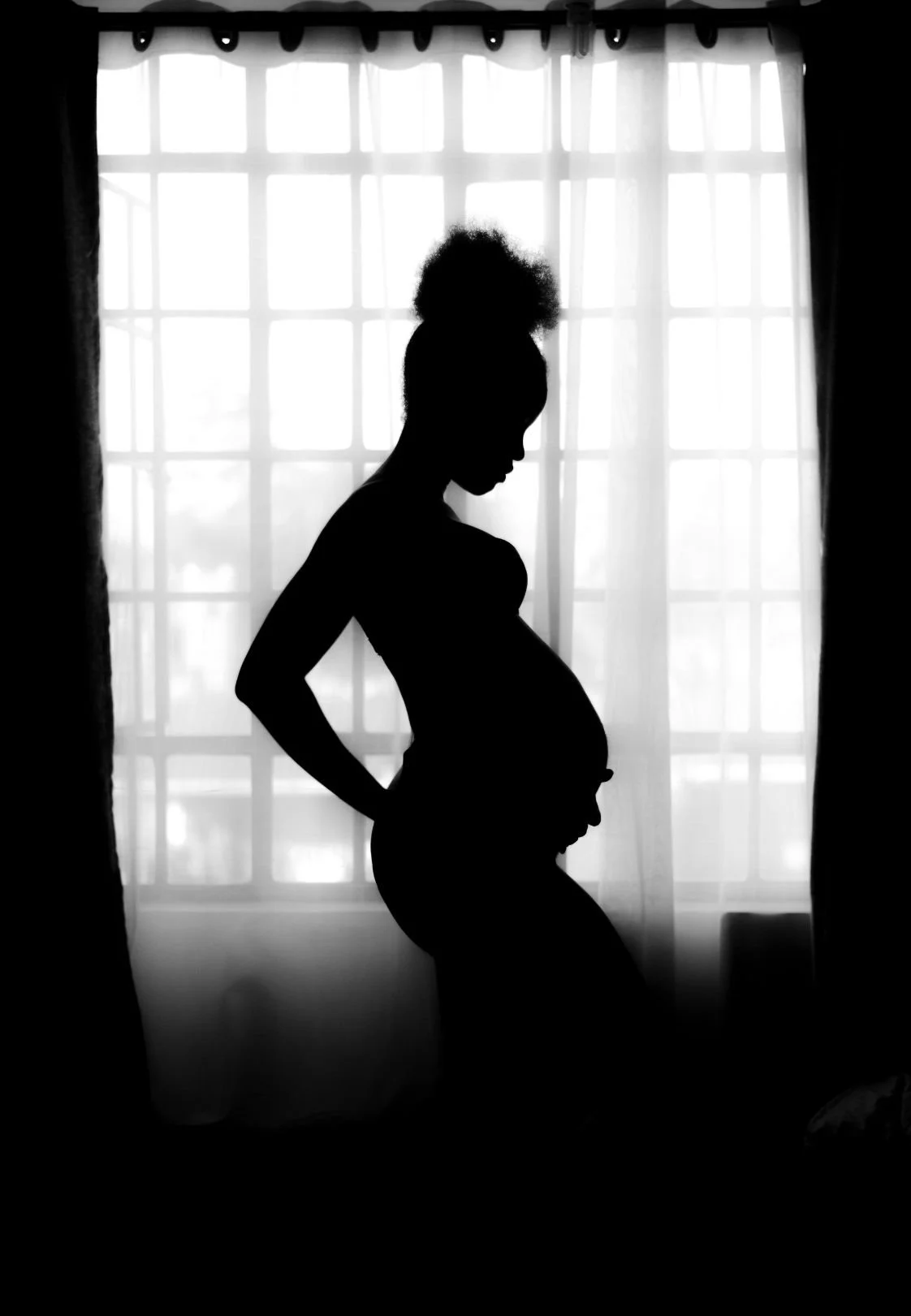 Silhouette of a pregnant woman standing in front of a window with sheer curtains, with her hands on her hips and her belly visible.
