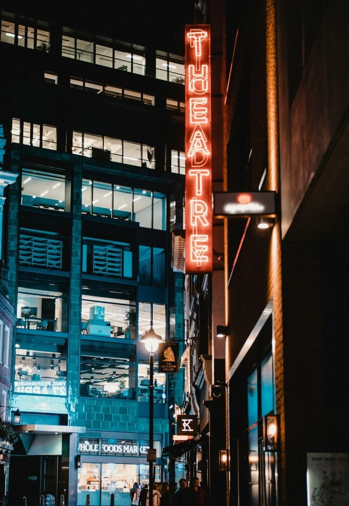 Nighttime city street scene with illuminated neon signs, including a vertical red sign reading 'TEA DT R TU' and other signs for stores and restaurants, with pedestrians walking along the sidewalk.