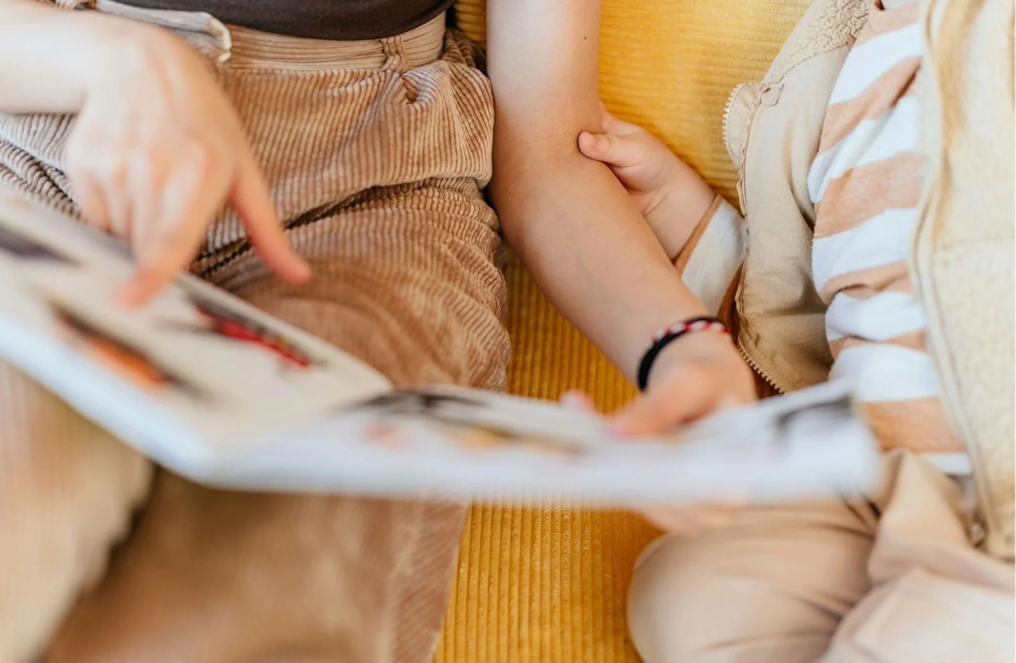Close-up of children holding hands, with one child wearing a striped shirt and the other a brown corduroy skirt, lying on a yellow textured surface.