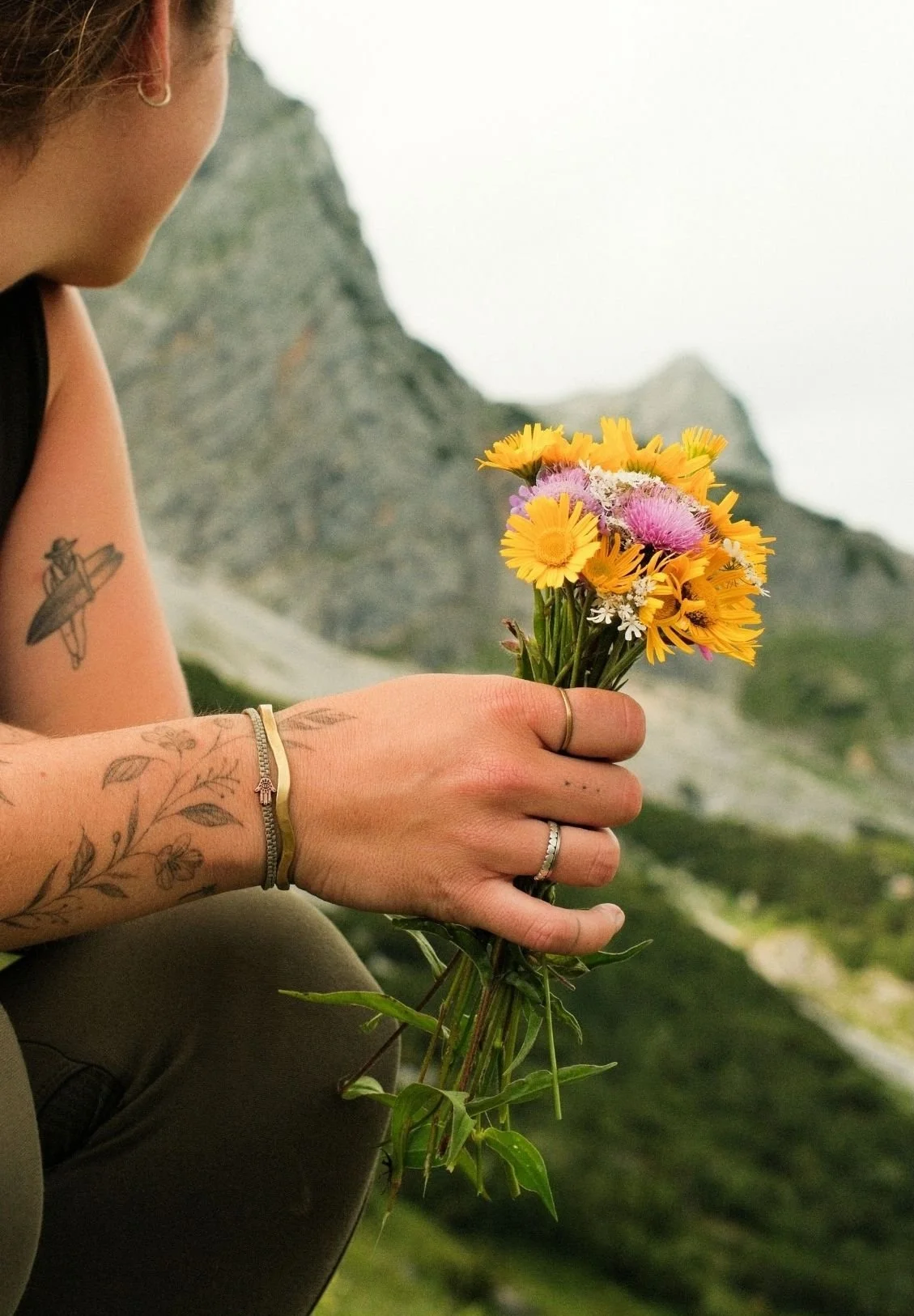 A person holding a small bouquet of colorful wildflowers outdoors with a mountain in the background.