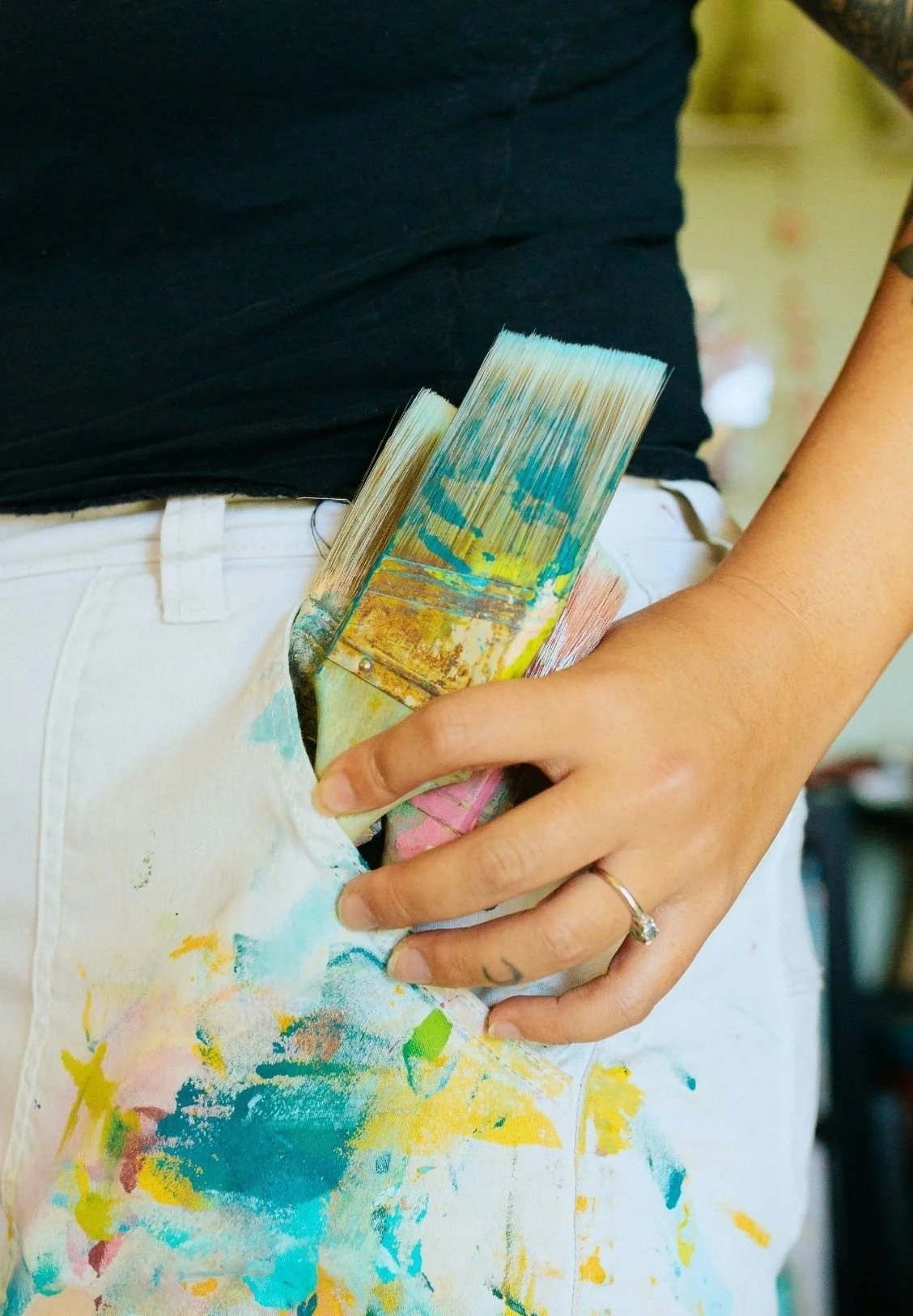 Person wearing white pants with colorful paint splatters holding a paintbrush in their pocket.