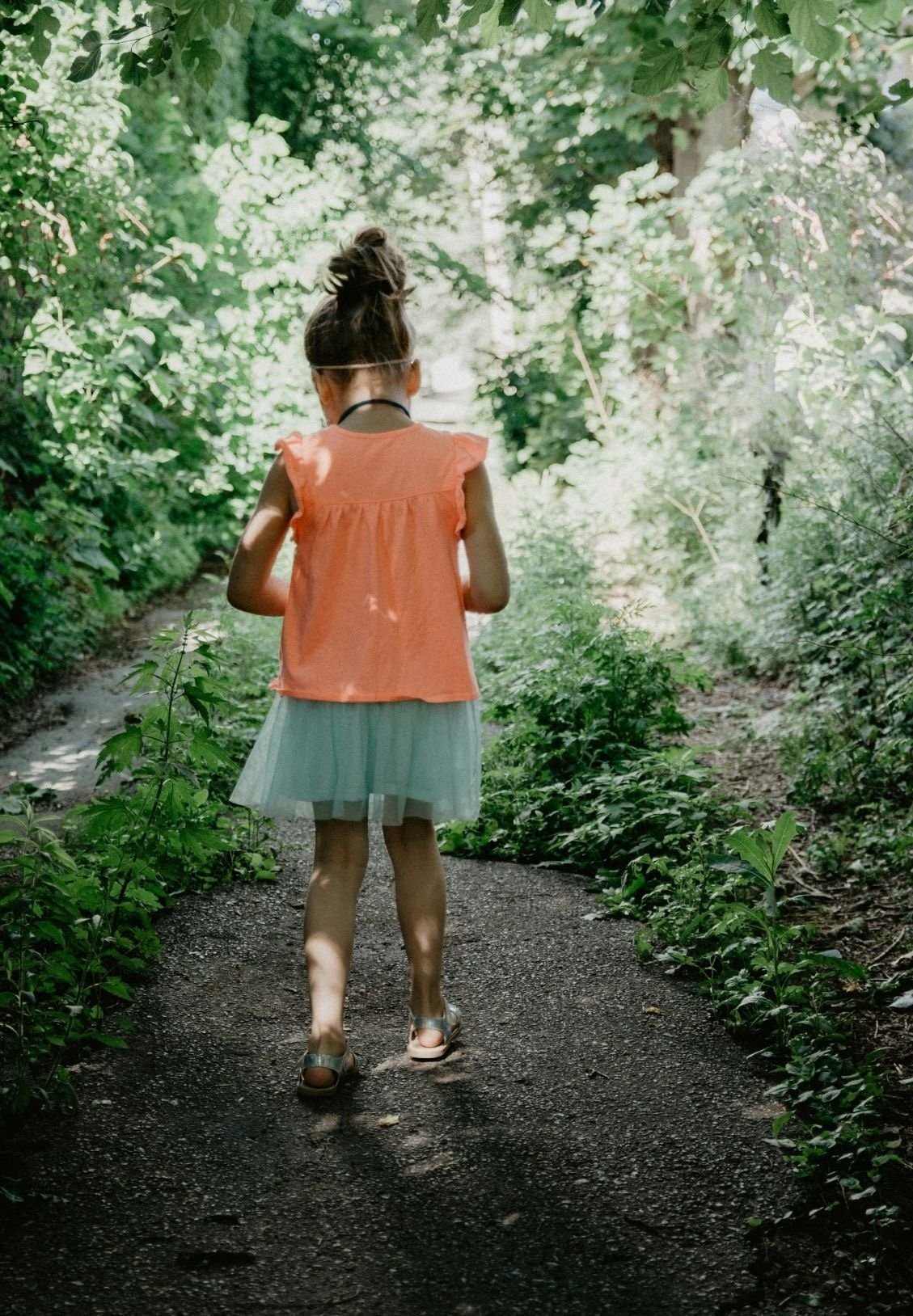 A girl wearing a peach top and a light green skirt walking along a narrow dirt trail through a lush green forest.