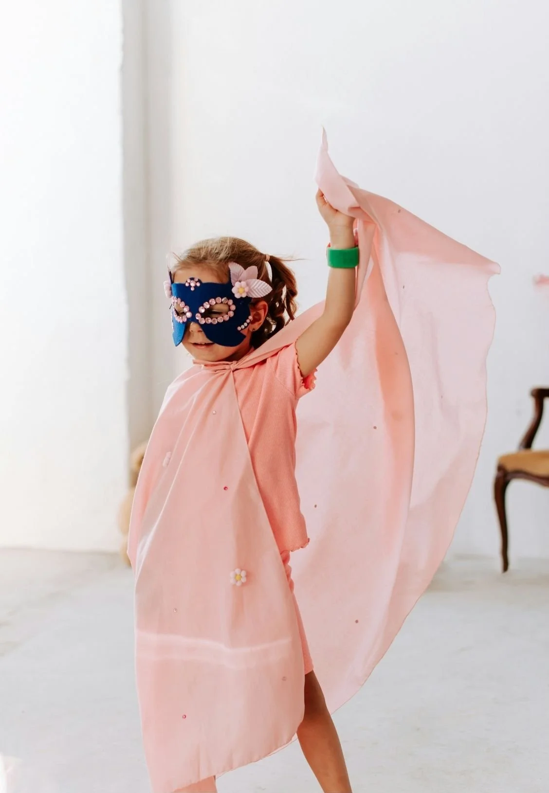 A smiling young girl wearing a blue mask with flower decorations, a pink cape, and matching dress, waving a pink cloth in a bright room with minimal furniture.