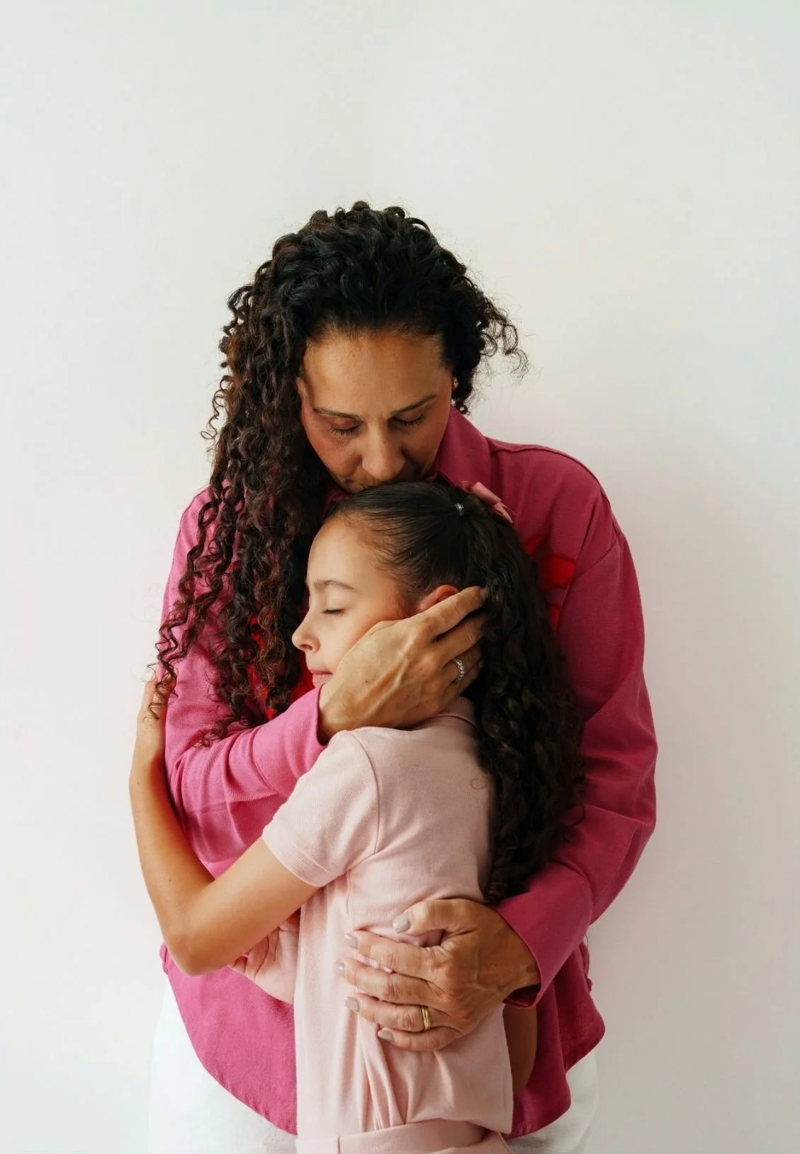 A woman and a young girl embrace each other, with the woman gently touching the girl's face and holding her in a warm hug against a plain white background.