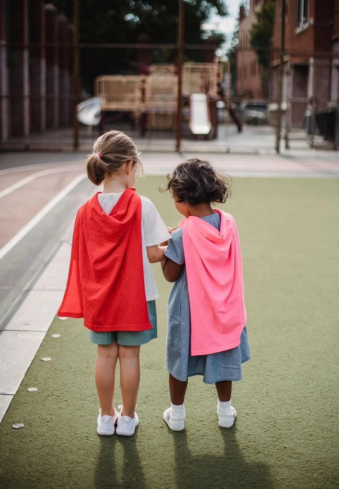 Two young girls standing on a sports court with artificial grass, wearing hooded capes, one red and one pink, and looking at something together.