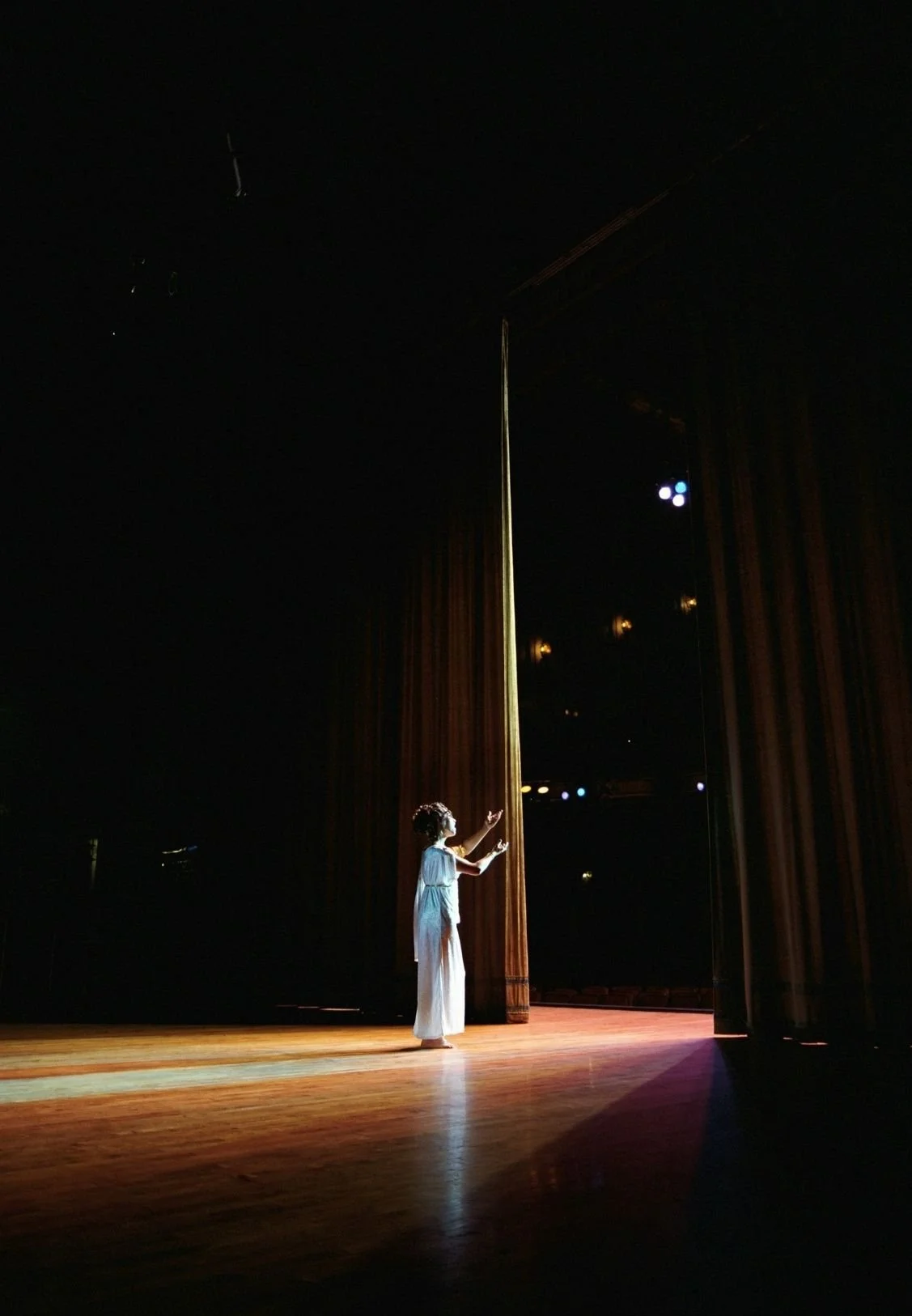 A woman standing on stage during a performance, reaching towards a tall pole or fabric hanging from the ceiling, with stage lights illuminating her and the stage floor, but the background is dark.