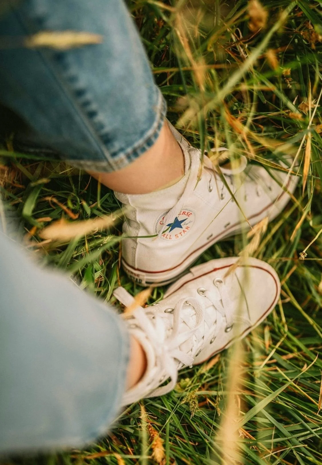 Close-up of white Converse All Star sneakers and denim jeans standing on grass.