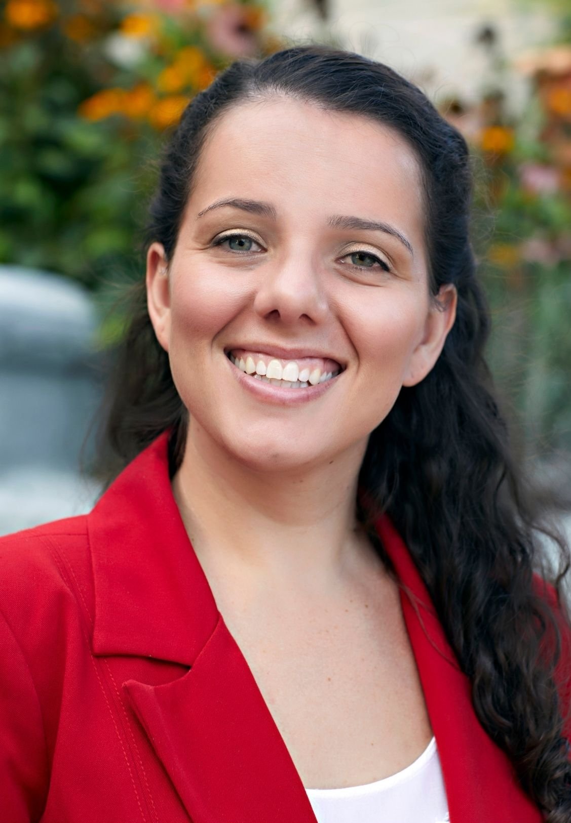 Ashley Martinez, Therapist in Lakewood, Colorado, with long dark hair, wearing a red blazer and a white top, outdoors with blurred greenery and flowers in the background.