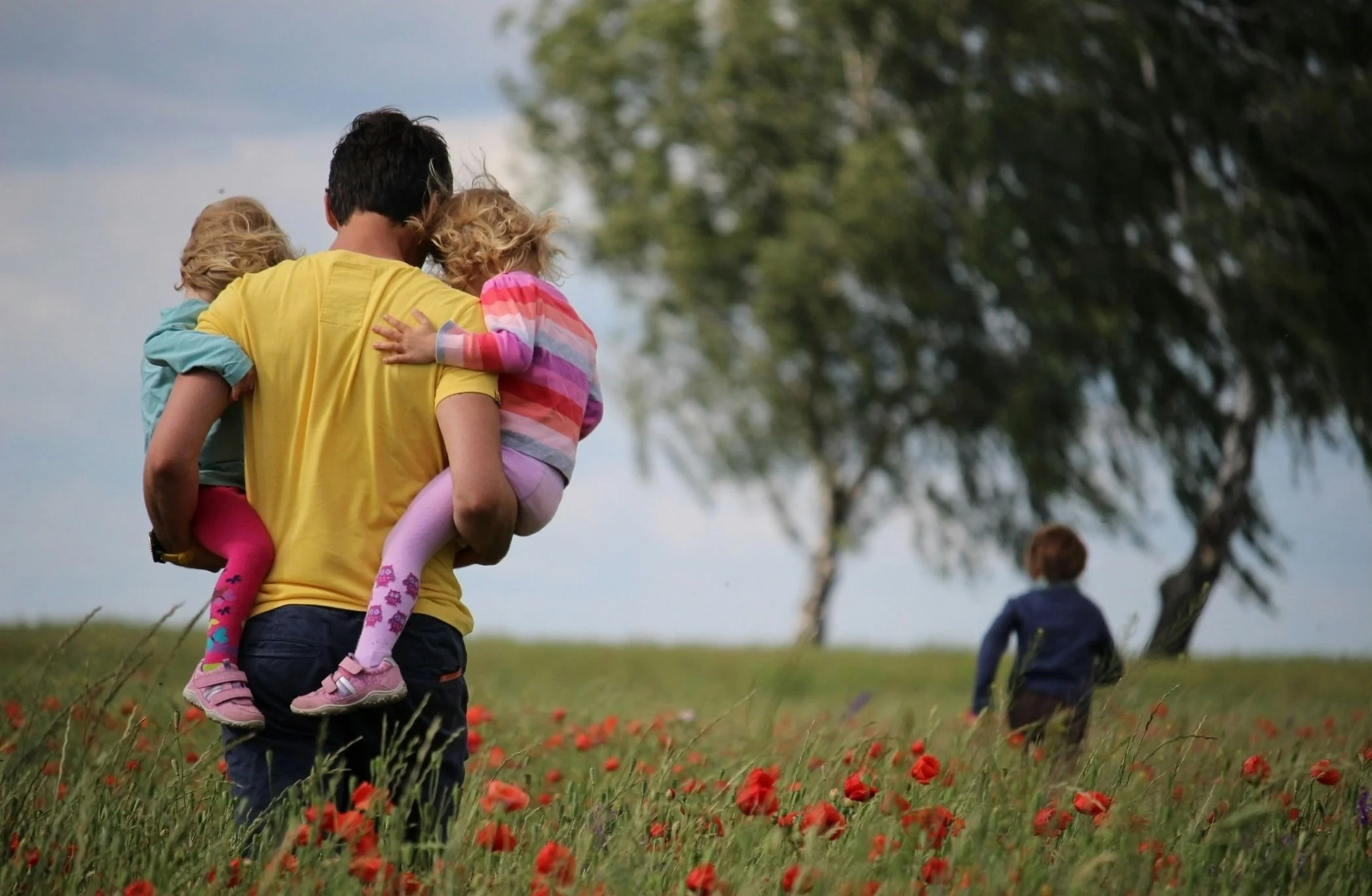 A man in a yellow shirt carrying two children in a grassy field with red flowers. One child is wearing pink leggings and a colorful striped top, while the other is wearing a blue jacket. In the background, there is a person with curly hair and a dark jacket, and trees under a cloudy sky.