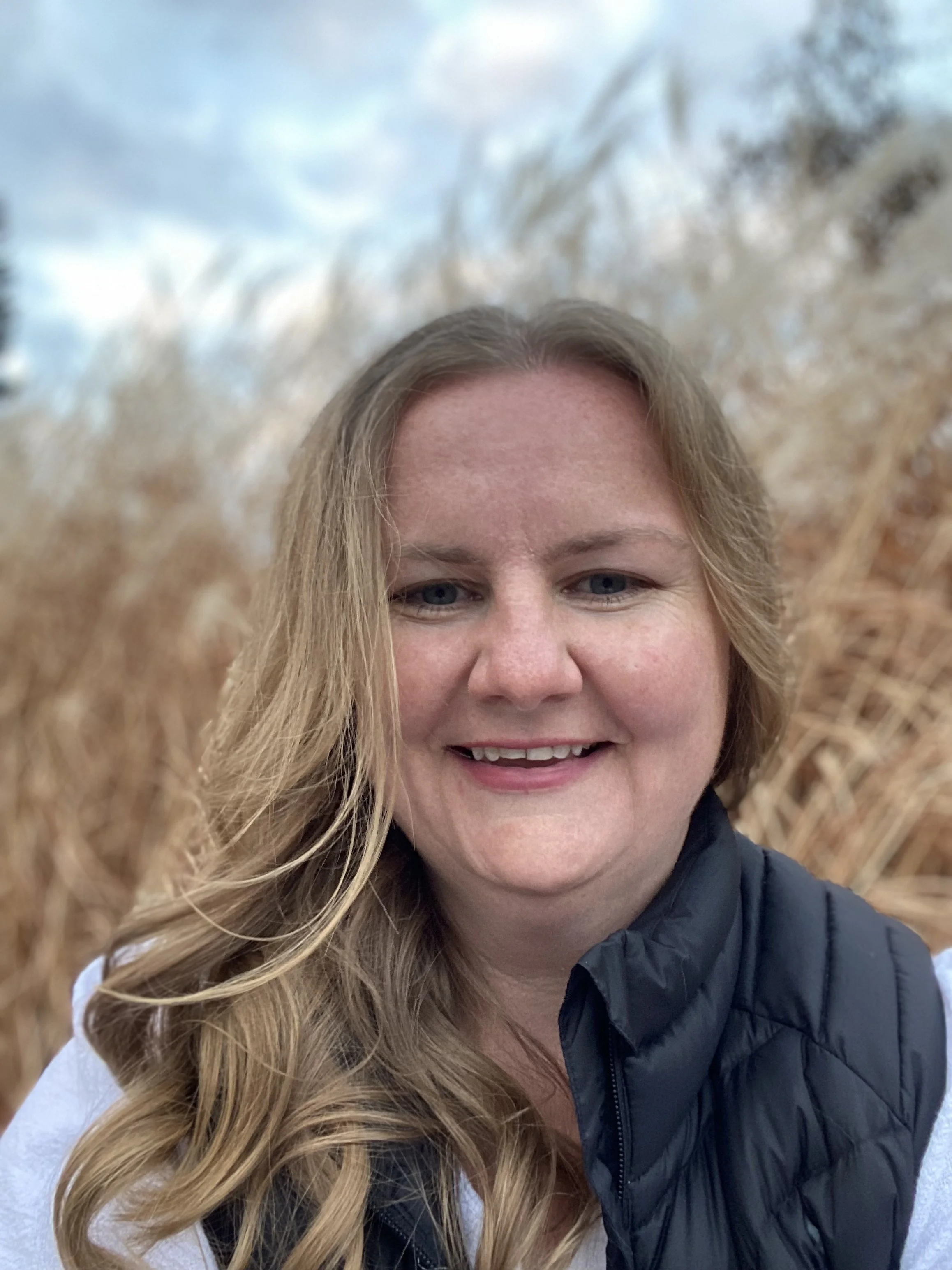 A smiling woman with long blonde wavy hair is outdoors with blurred trees and sky in the background, wearing a black jacket and white and black clothing.