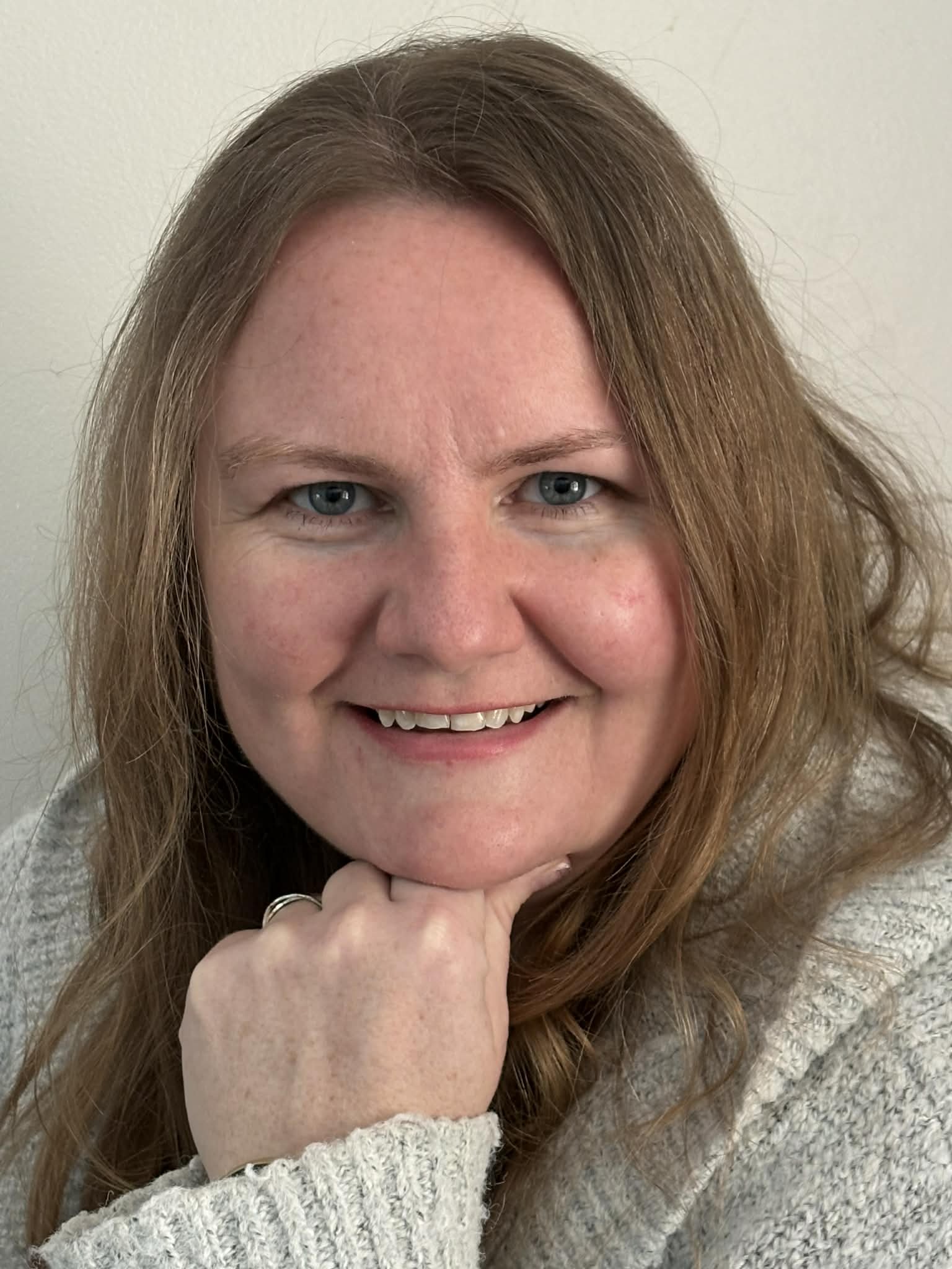 A woman with long, wavy red hair and blue eyes smiling at the camera, wearing a cozy light gray sweater while resting her chin on her hand.
