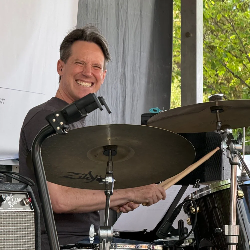 A man smiling and playing drums outdoors with greenery in the background.