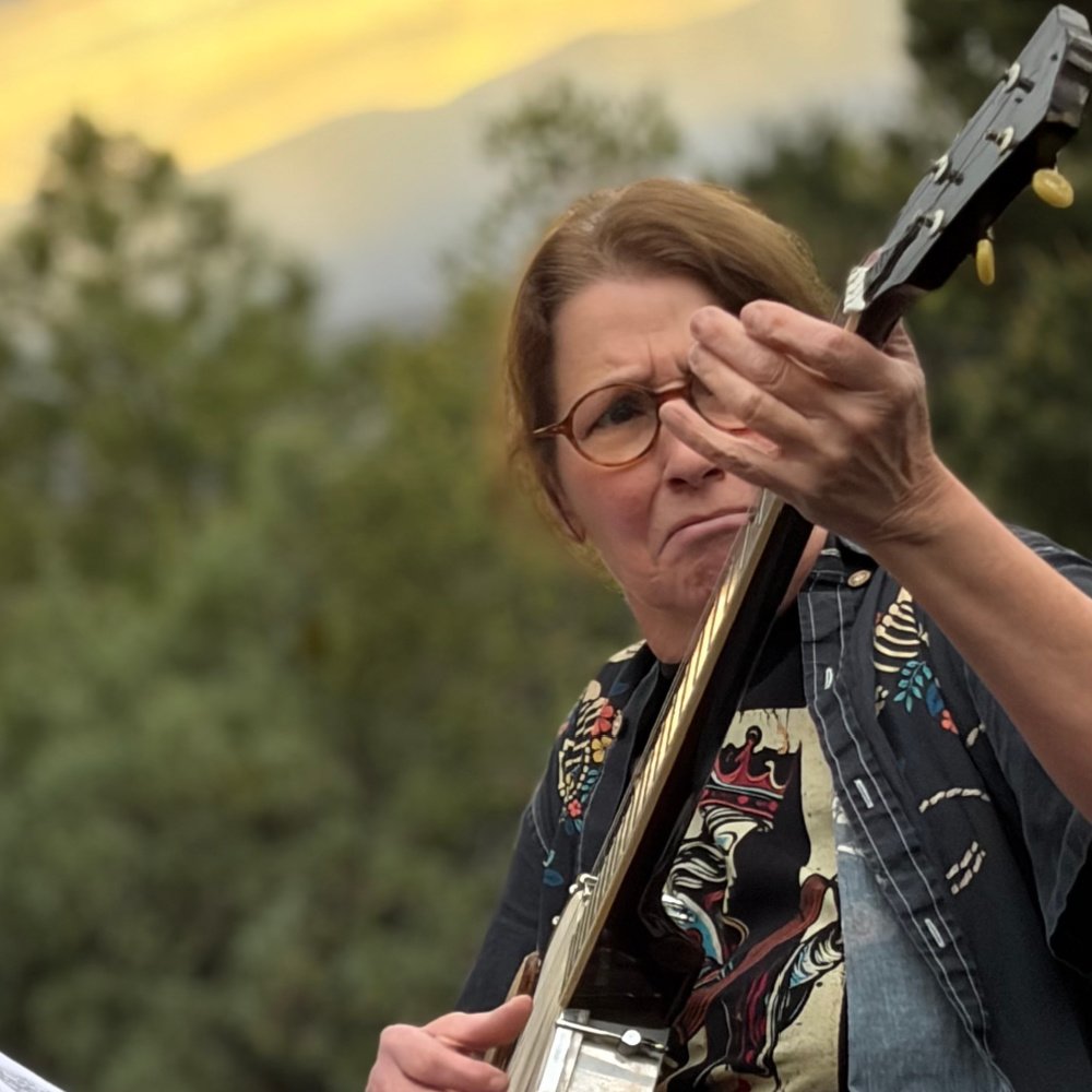 Woman with glasses playing an acoustic guitar outdoors during sunset, in a natural setting with trees in the background.