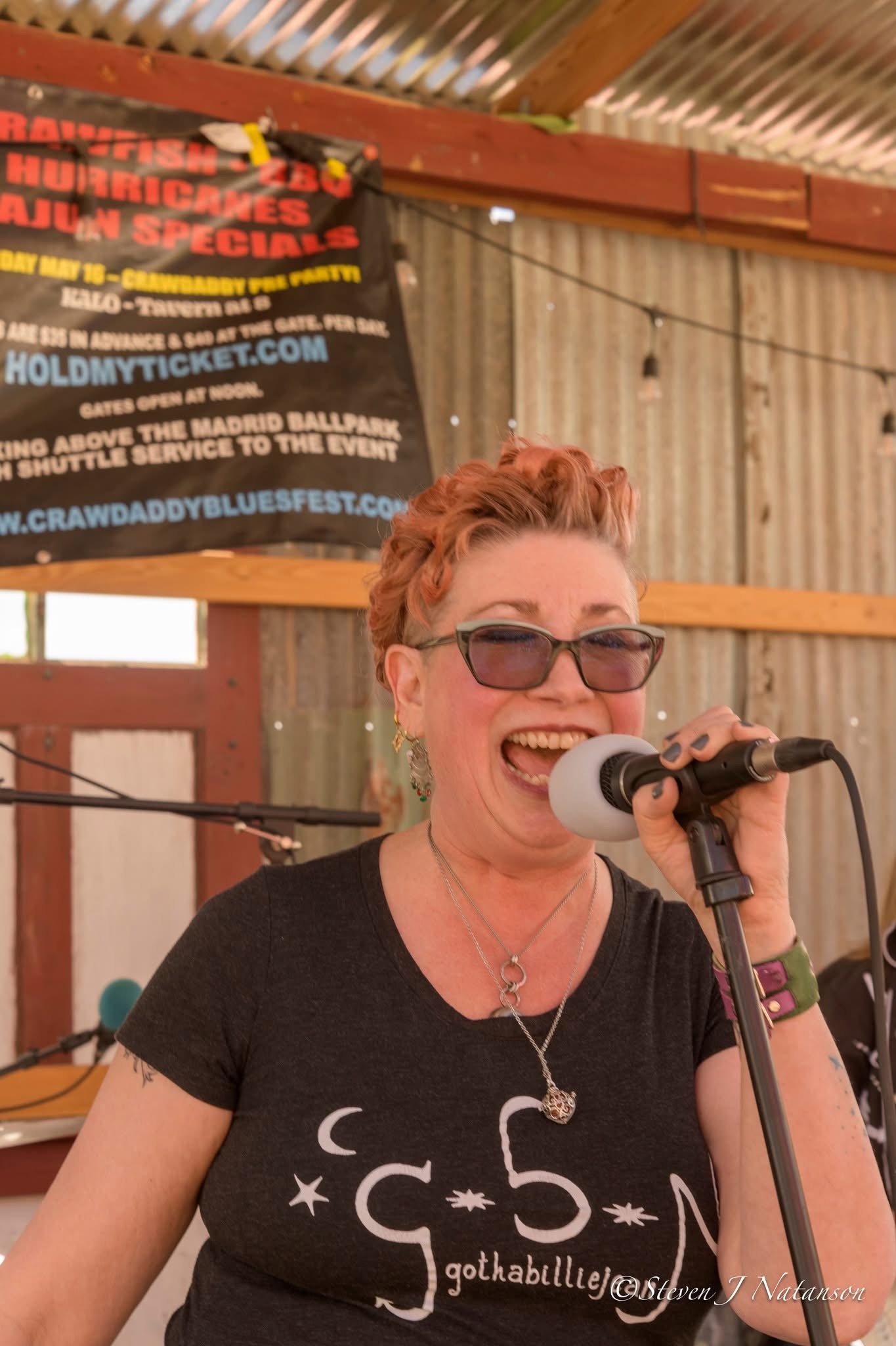 A woman with short curly hair and glasses singing into a microphone at an indoor event. She is wearing a black shirt with a moon and star design and the words "gothabilleje". In the background, there is a banner advertising the Crawdaddy Blues Festival, and the setting features wooden walls and ceiling beams.