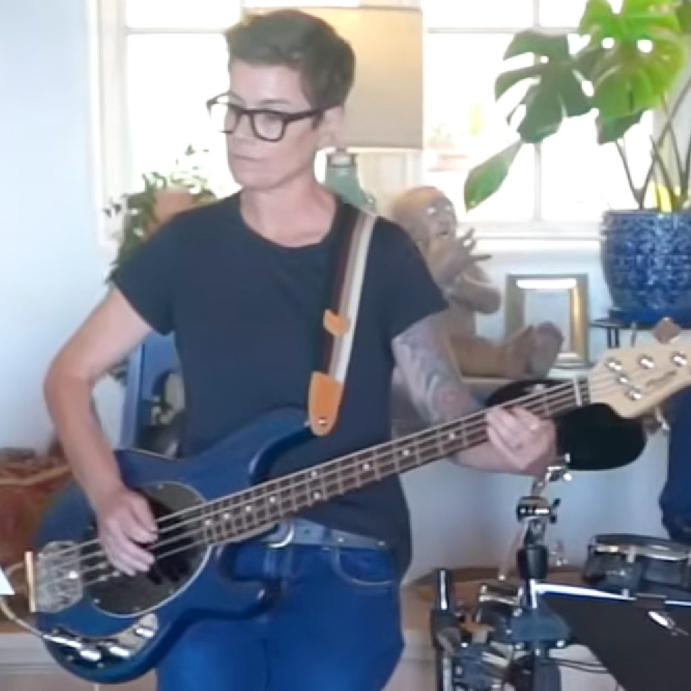A person with short dark hair, glasses, and a black T-shirt playing a blue electric bass guitar indoors surrounded by plants and musical equipment.