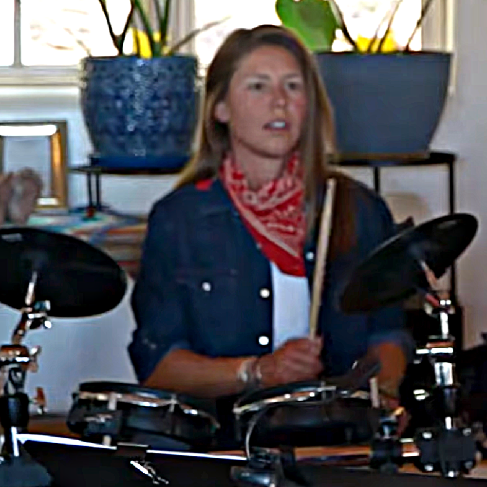 A woman with long brown hair, wearing a red bandana around her neck, playing electronic drums indoors. Behind her are large blue planters with green plants and a window.