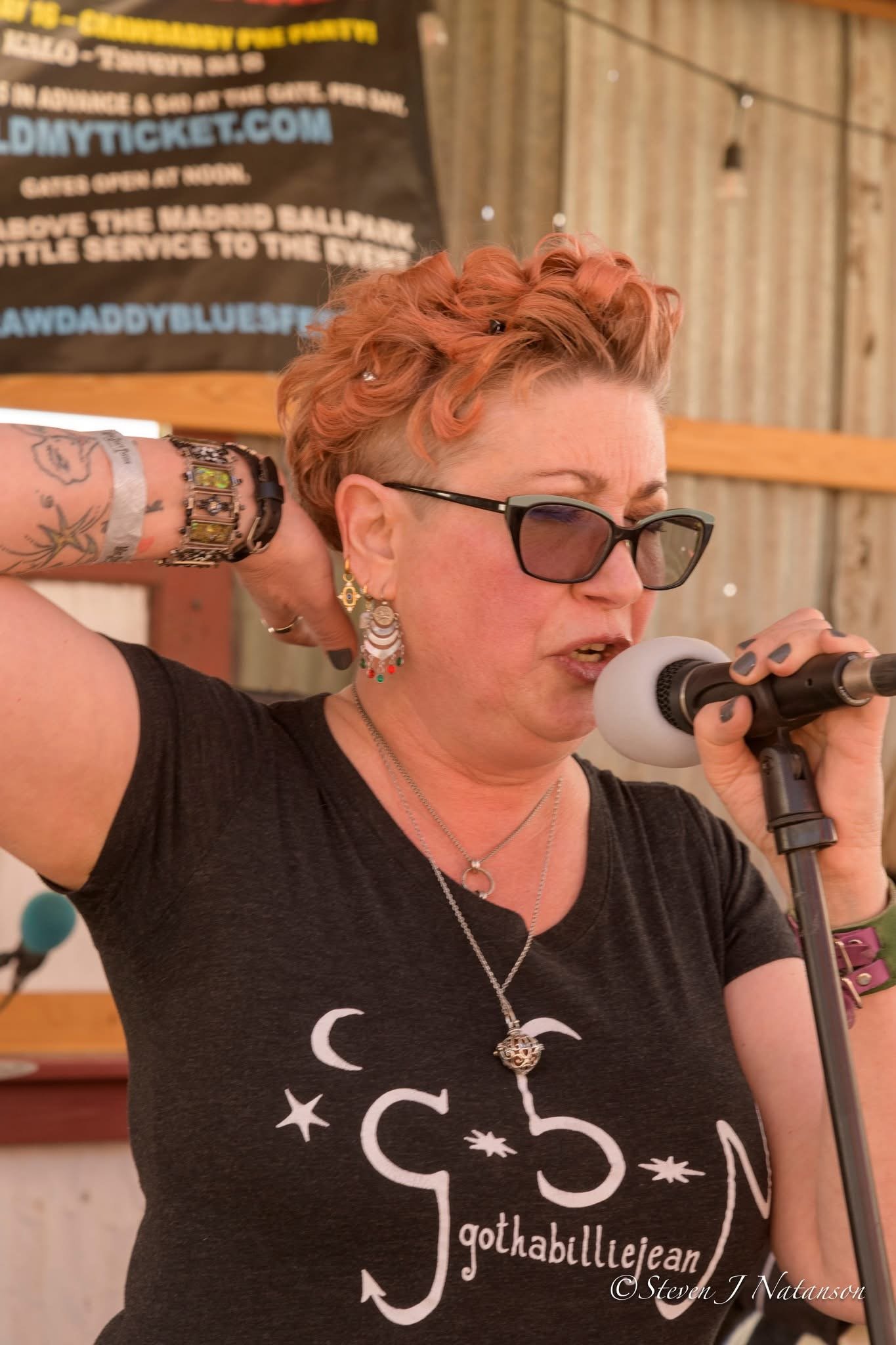 A woman with short curly red hair and sunglasses singing into a microphone at an event. She is wearing a black T-shirt with white celestial drawings and the words 'gothabulliejean'. She has tattoos on her arm and wears jewelry including earrings, a necklace, and a ring.