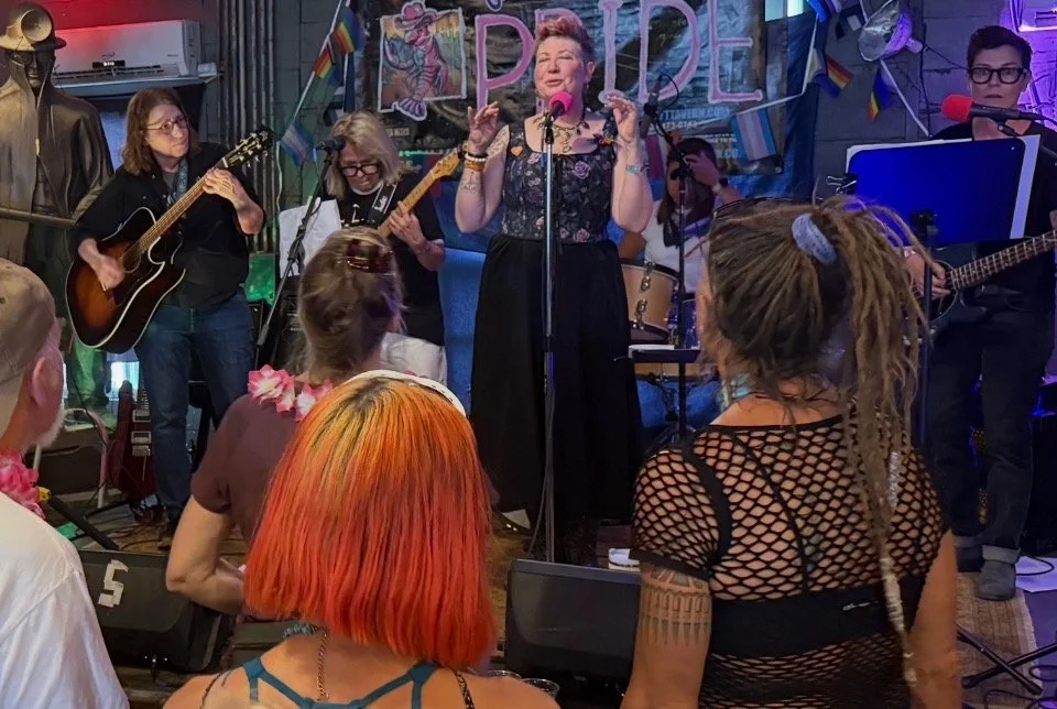A group of musicians and a singer performing at a pride event, with an audience watching. The background has pride flags and a sign that says 'PRIDE'.
