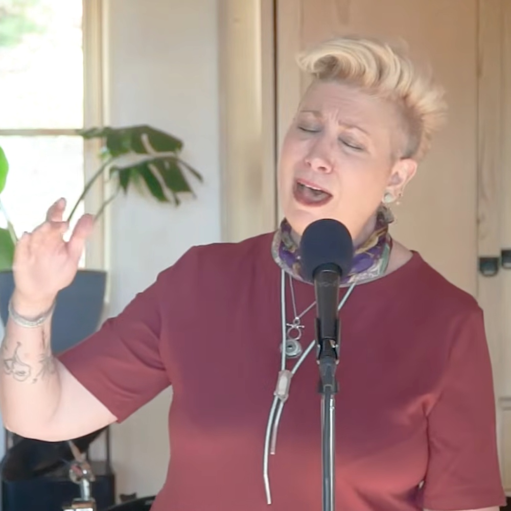 A person with short blonde hair singing passionately into a microphone, wearing a maroon shirt and a colorful scarf, standing indoors near a window and a potted plant.