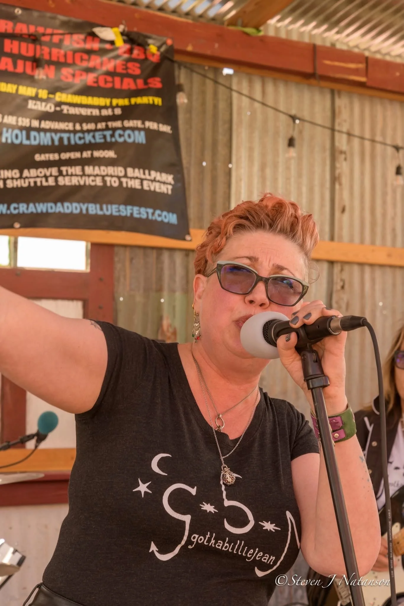 A woman with short red hair and glasses singing into a microphone at an event. She is wearing a black T-shirt with a moon and star design and the text "gothabilliejjean" printed on it. Behind her, there is a wooden wall and a colorful banner advertising a blues festival.