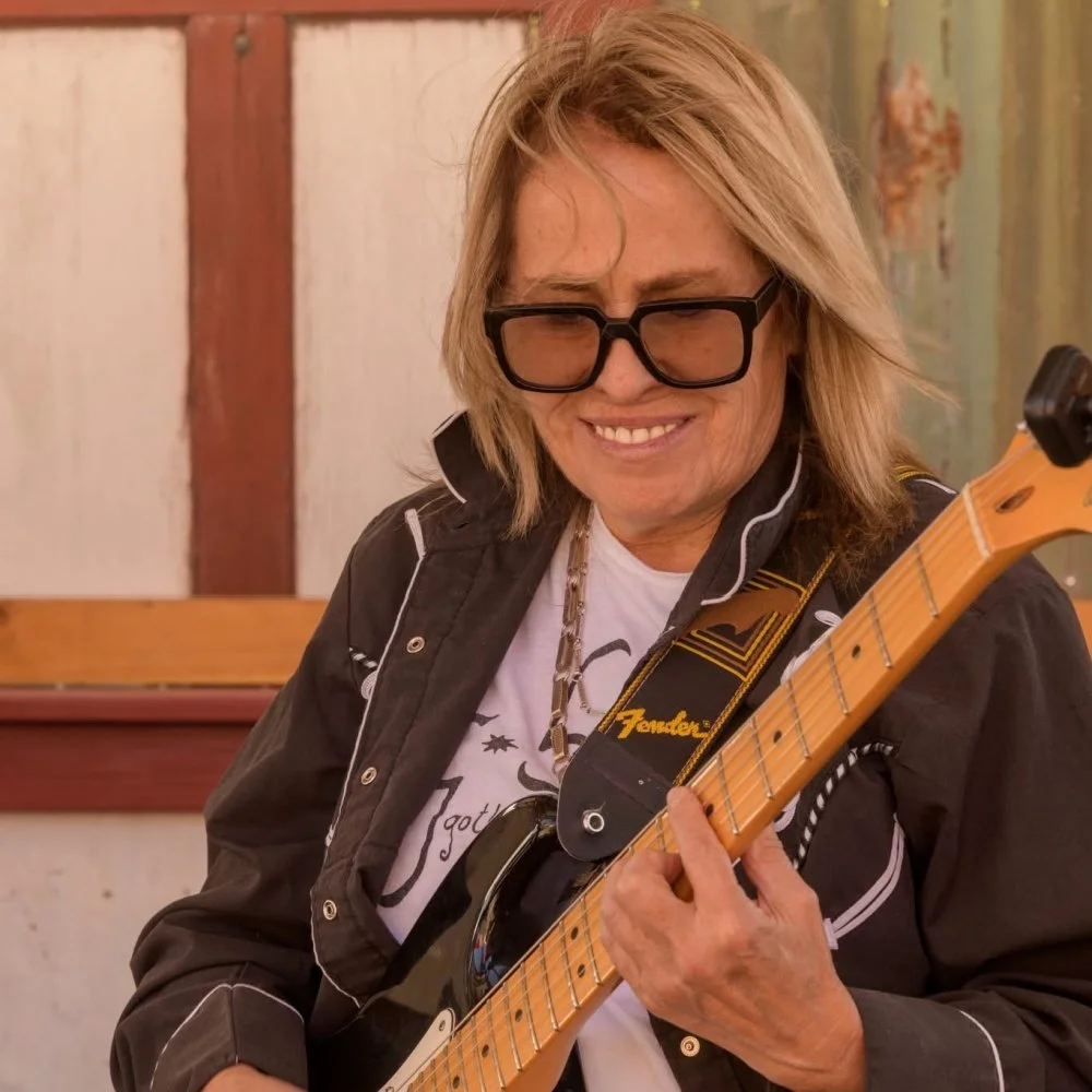 A woman with blonde hair, wearing glasses, smiling and playing an electric guitar.