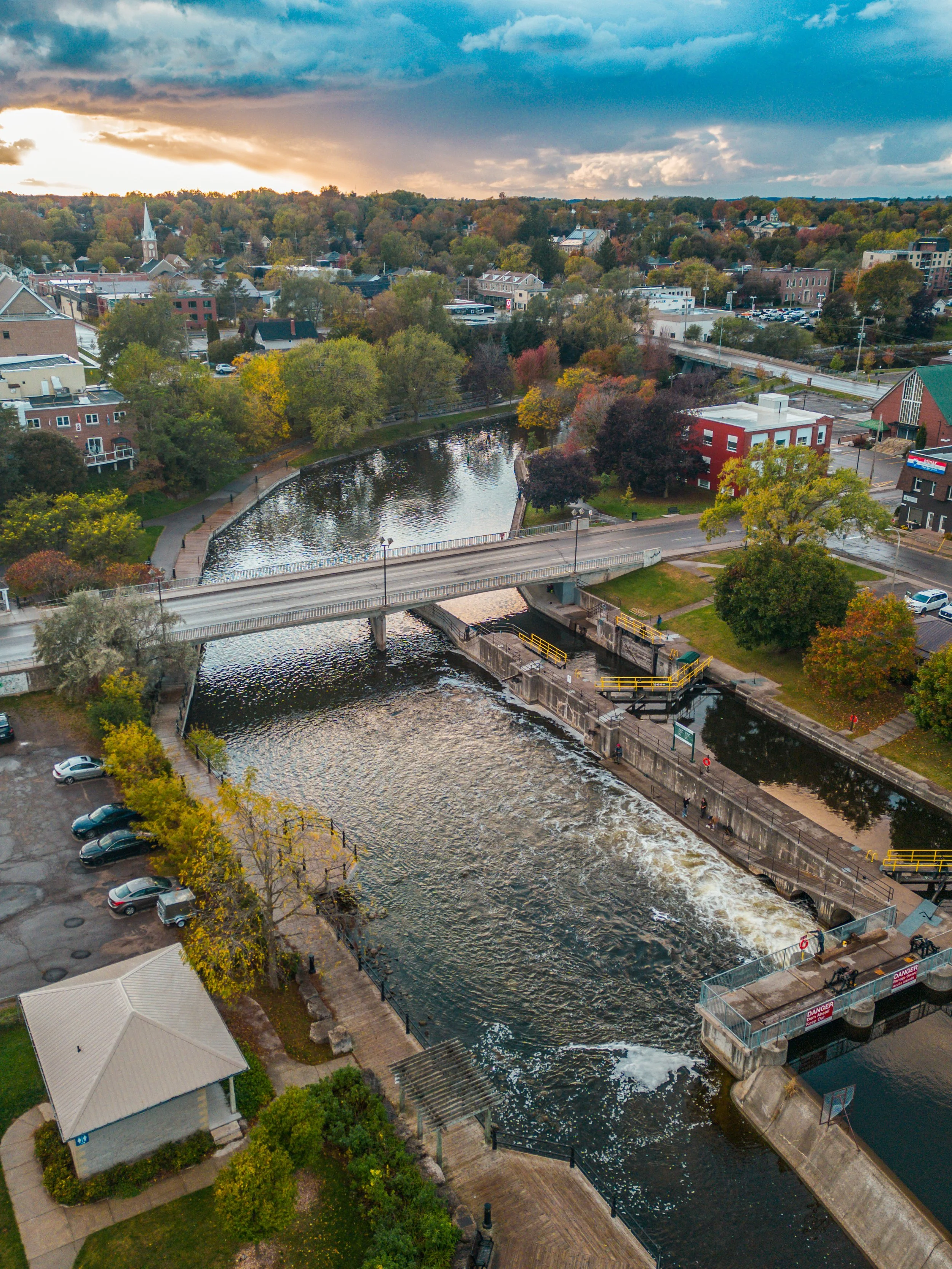 Aerial view of a small town with a river running through it, surrounded by trees with fall foliage, a bridge crossing the river, and various buildings and parking lots.