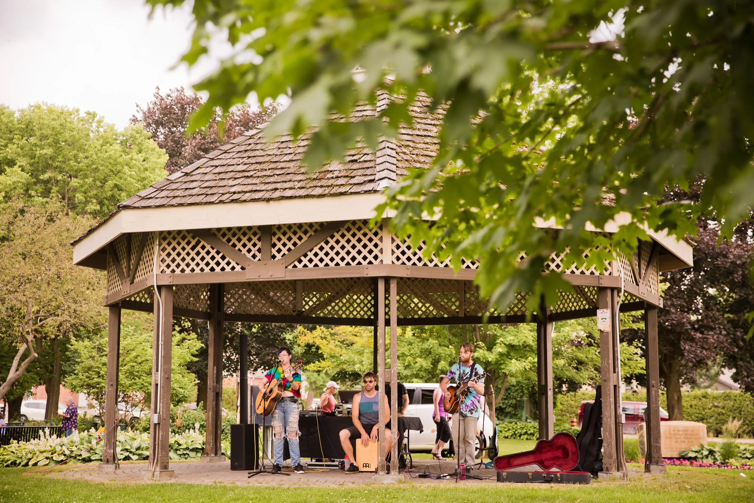 A small outdoor stage with musicians performing, surrounded by trees and greenery, with a few people in the background.