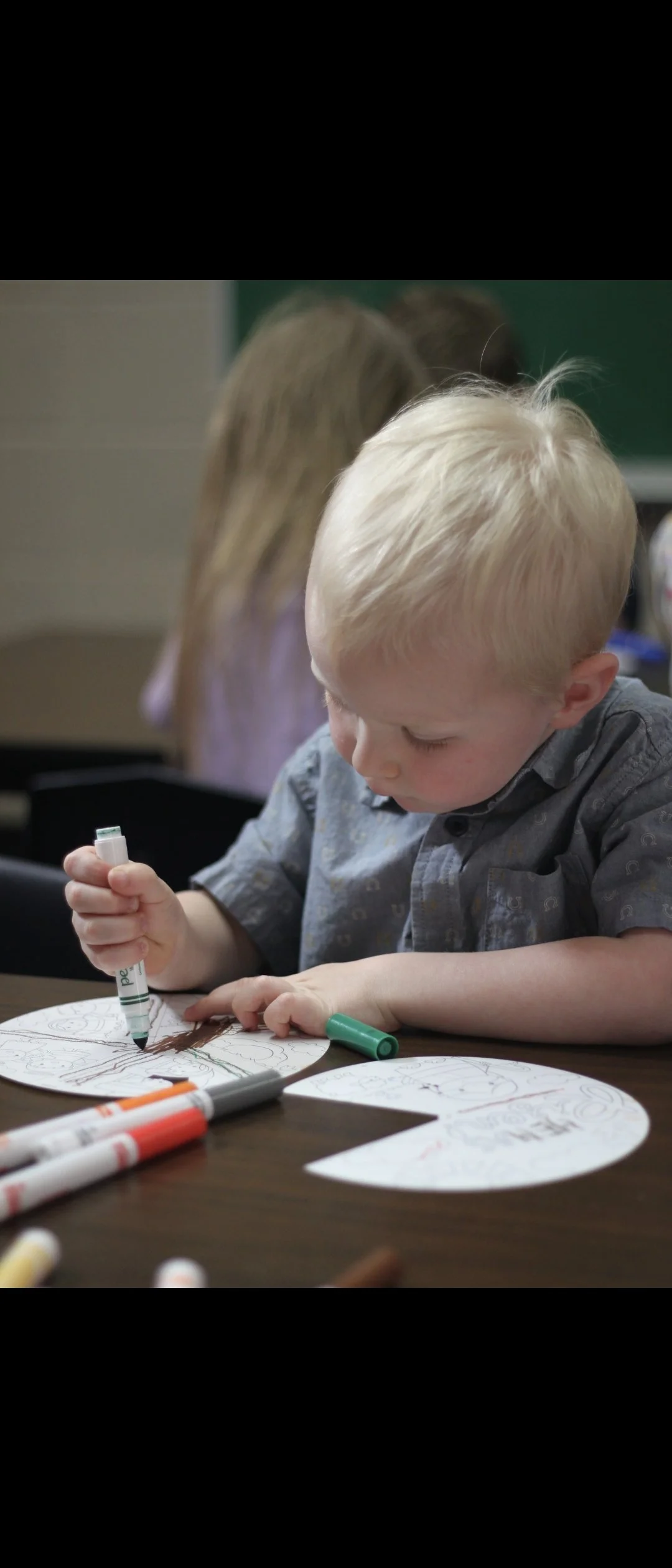 Young boy coloring with markers at a school desk, with adults in the background.