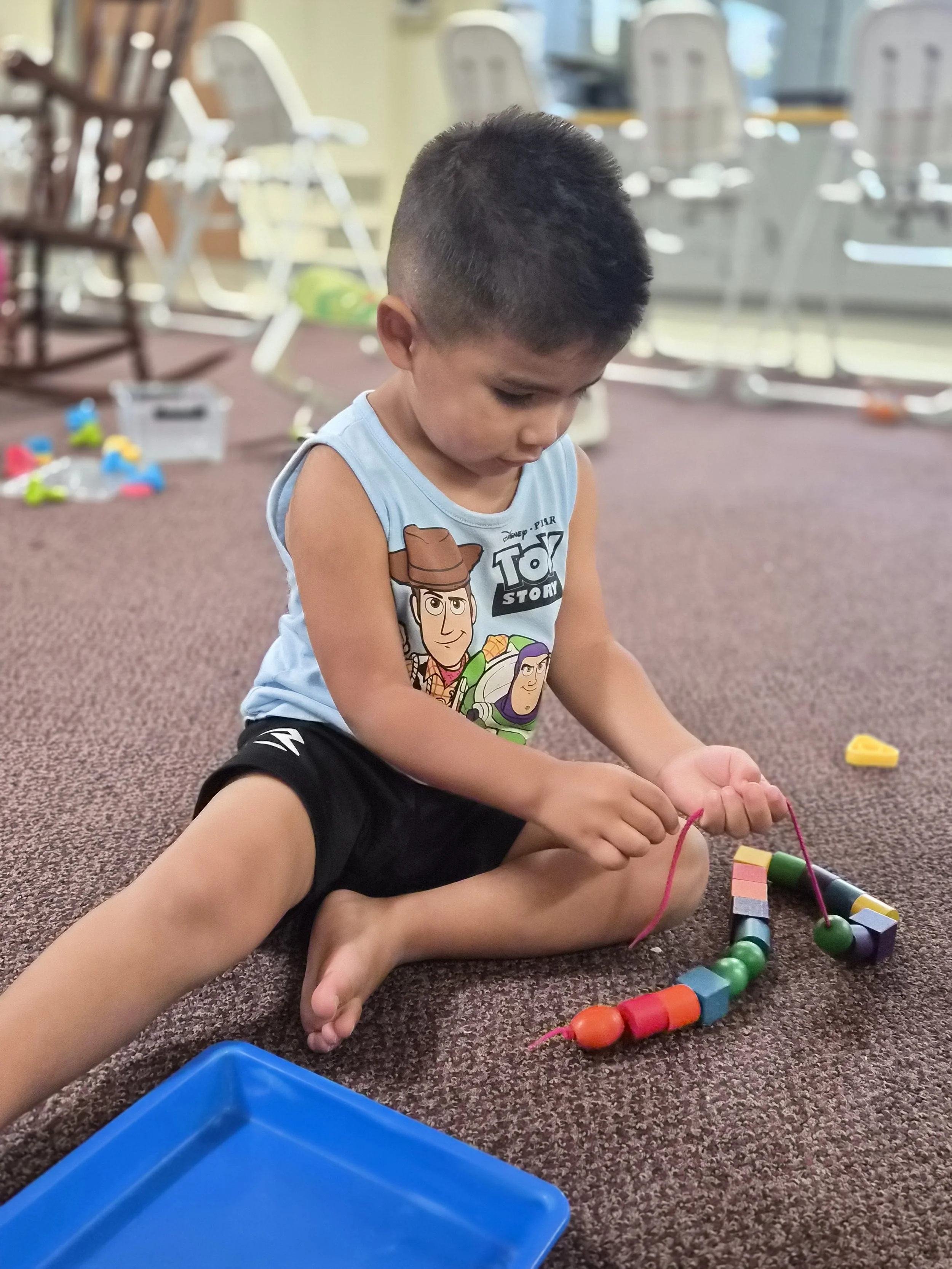 A young boy sitting on a brown carpeted floor playing with colorful wooden beads and string, with a blue plastic container nearby and chairs and windows in the background.