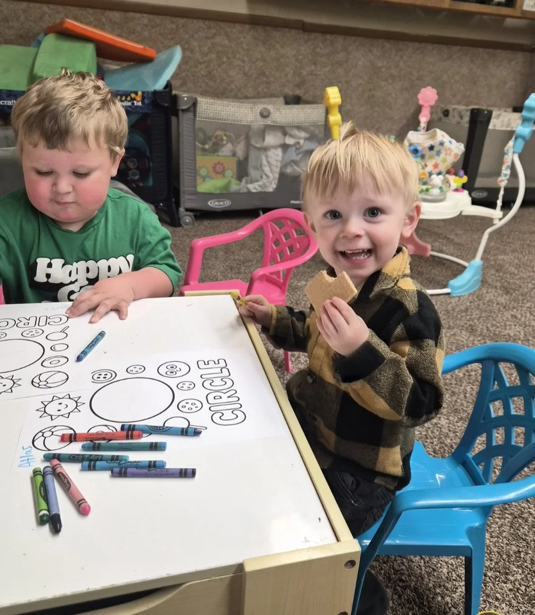 Two young boys sitting at a small table in a playroom. One boy in a green shirt is looking down at his drawing, while the other boy in a plaid shirt is smiling and holding a piece of cereal. The table has some colorful crayons and a printed coloring sheet with circles and the word 'CIRCLE'. In the background, there are toys, a playpen, and activity center.