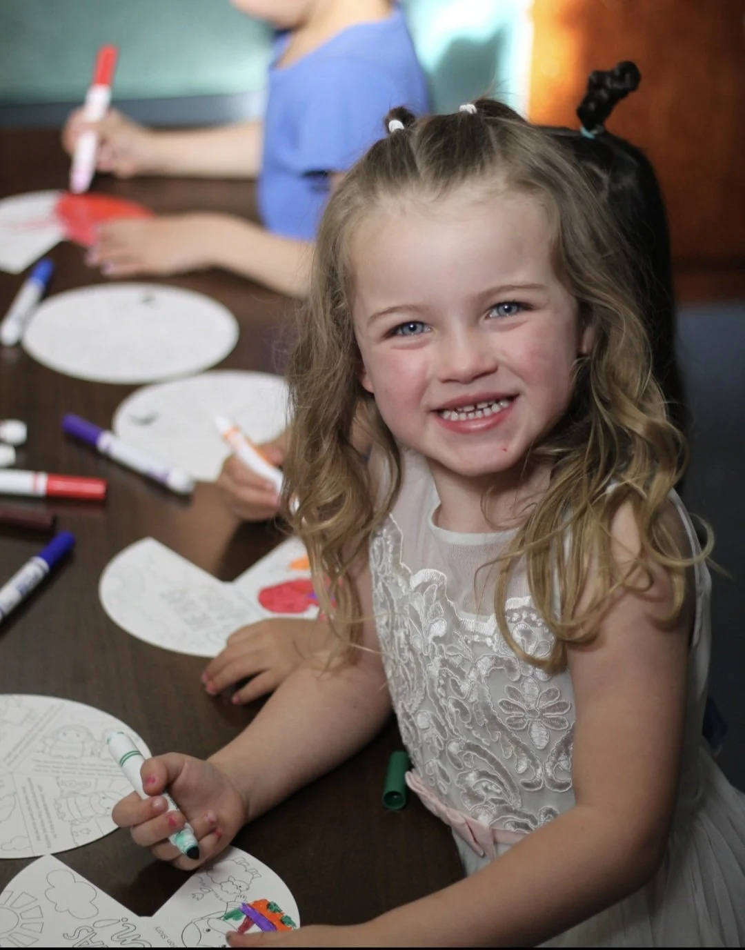 Young girl with curly blonde hair and blue eyes smiling at camera, sitting at a table with coloring pages and markers, with two other children in the background also coloring.