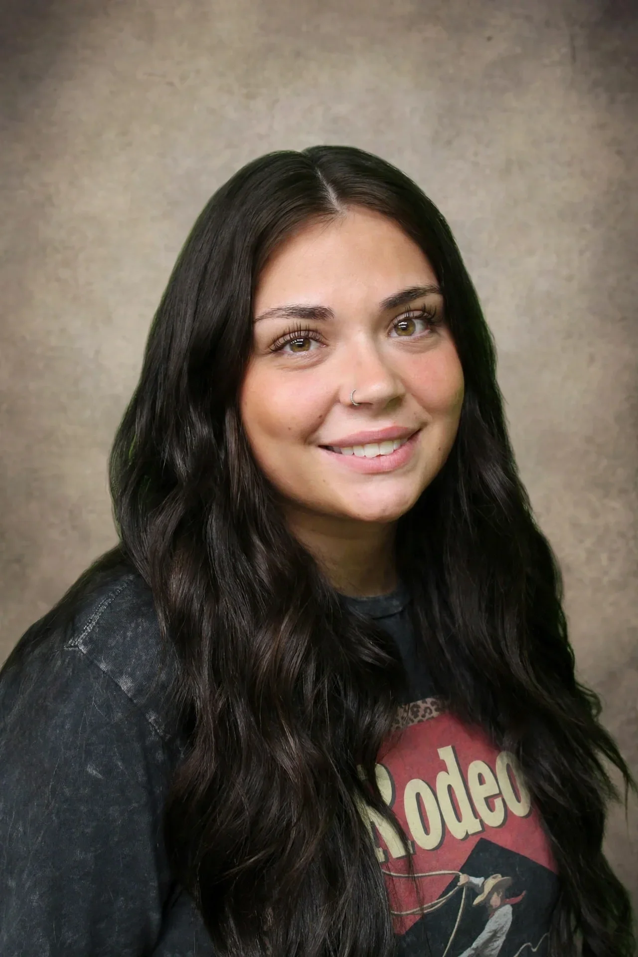 A young woman with long, wavy black hair, light brown eyes, and a nose ring, smiling, wearing a black jacket and a graphic t-shirt.