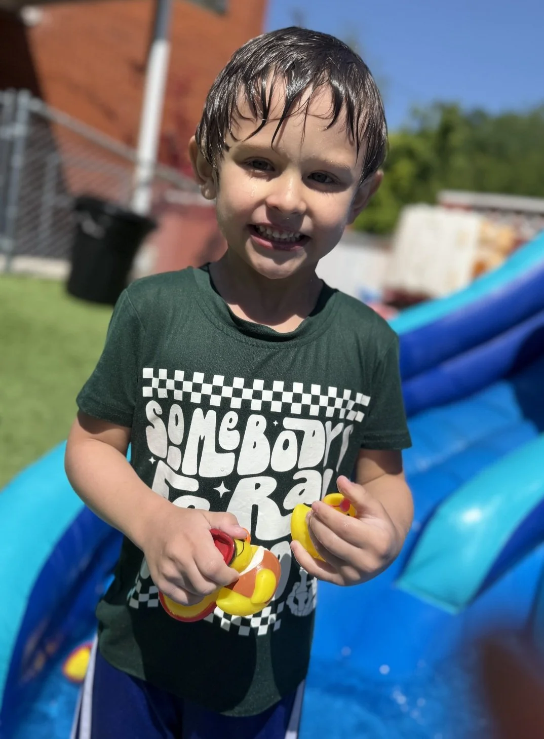 Young boy with wet hair smiling outdoors, holding yellow and red toy water guns, wearing a dark green t-shirt with white text, in a backyard with a blue inflatable pool.