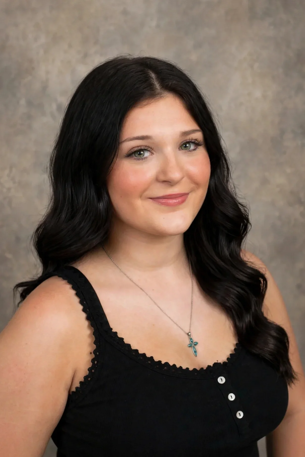 Portrait of a young woman with long, dark wavy hair, wearing a black sleeveless top with lace trim and a turquoise cross necklace, smiling against a neutral background.
