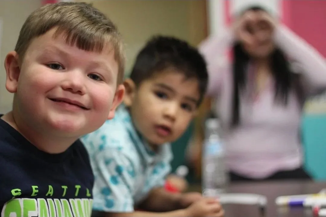 Three children in a classroom, one smiling at the camera, two others looking with surprised expressions; blurred background.