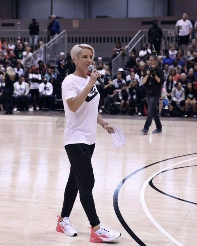 A woman standing on a basketball court holding a microphone and some papers, speaking to a large audience behind her.