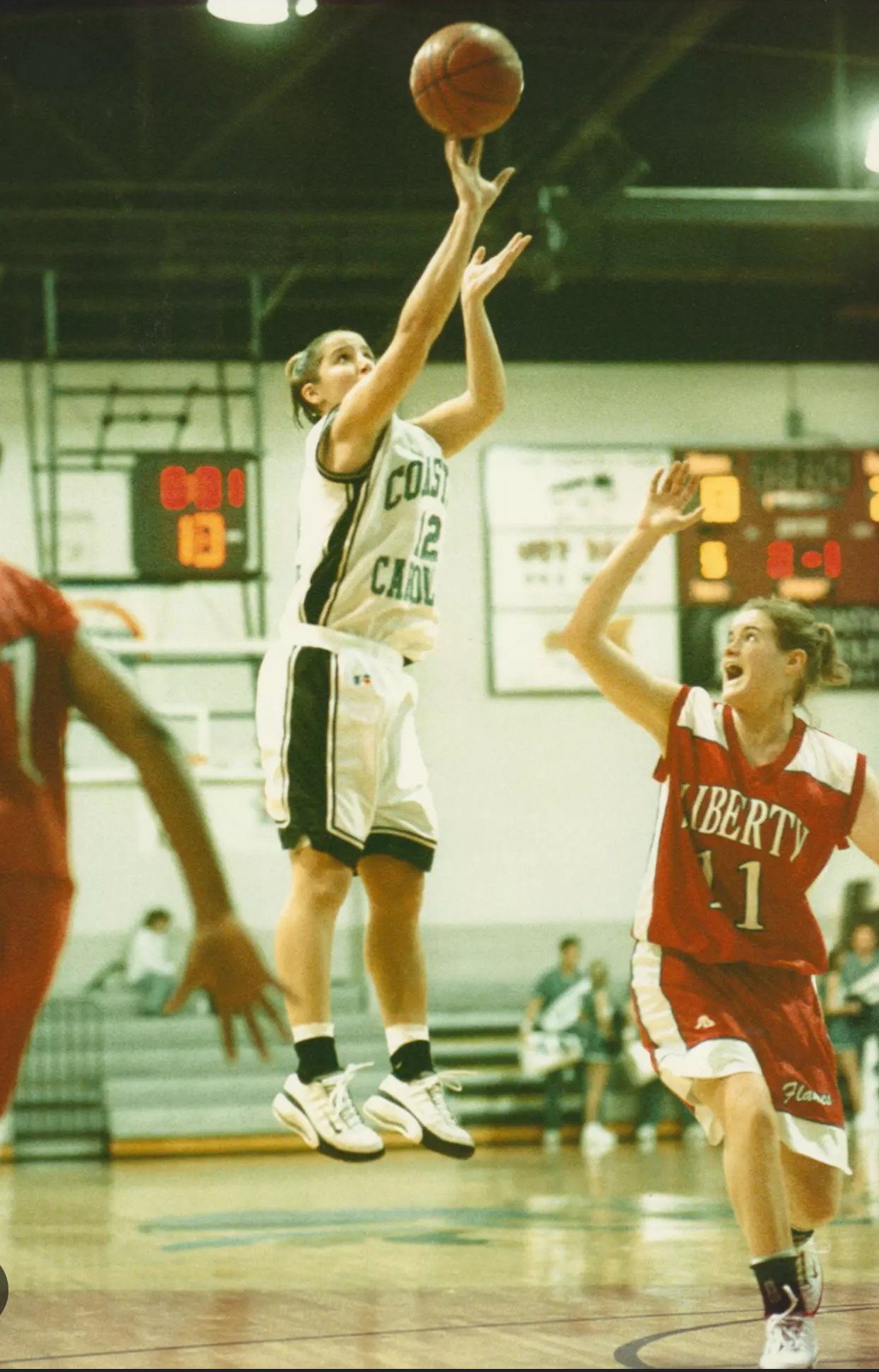 A woman in a white basketball uniform jumping to shoot the ball while another woman in a red uniform tries to block her during a game in a gymnasium.